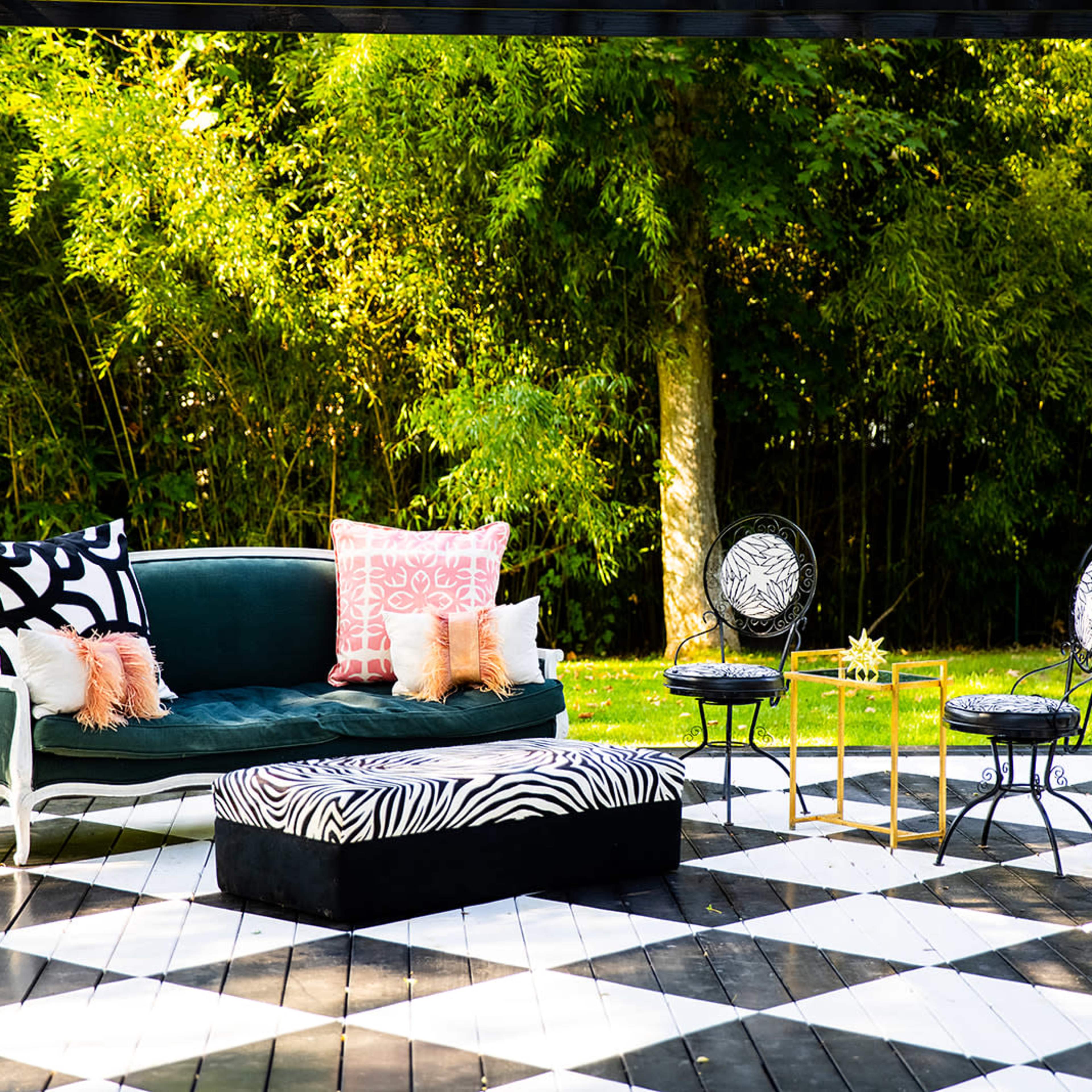 A stylish outdoor seating area features a green velvet sofa with decorative pillows, a black and white zebra-print ottoman, and elegant black chairs arranged on a checkered wooden deck surrounded by greenery.