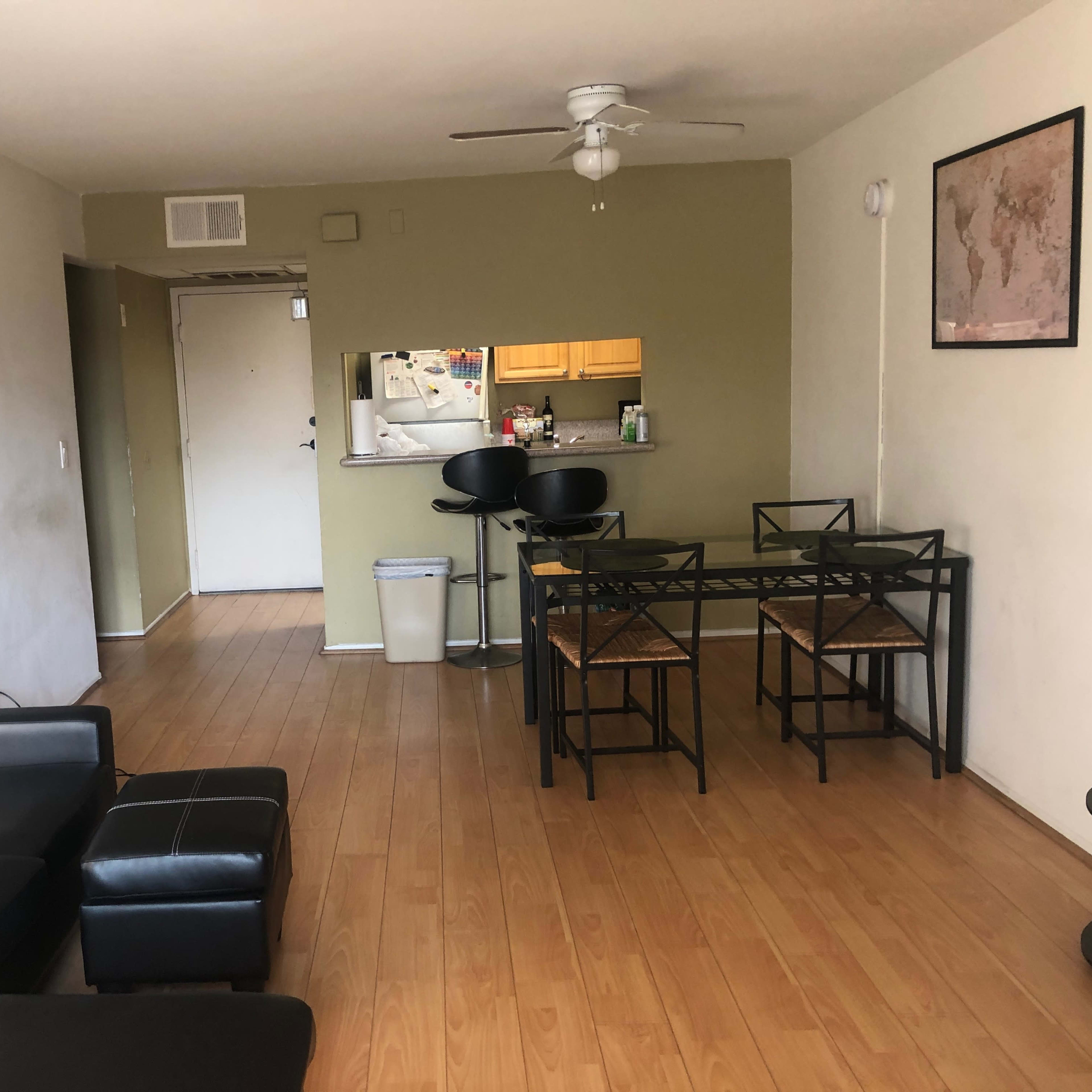 A living space with a black sofa, a matching ottoman, a glass dining table with four chairs, and a view of a kitchen area in the background.