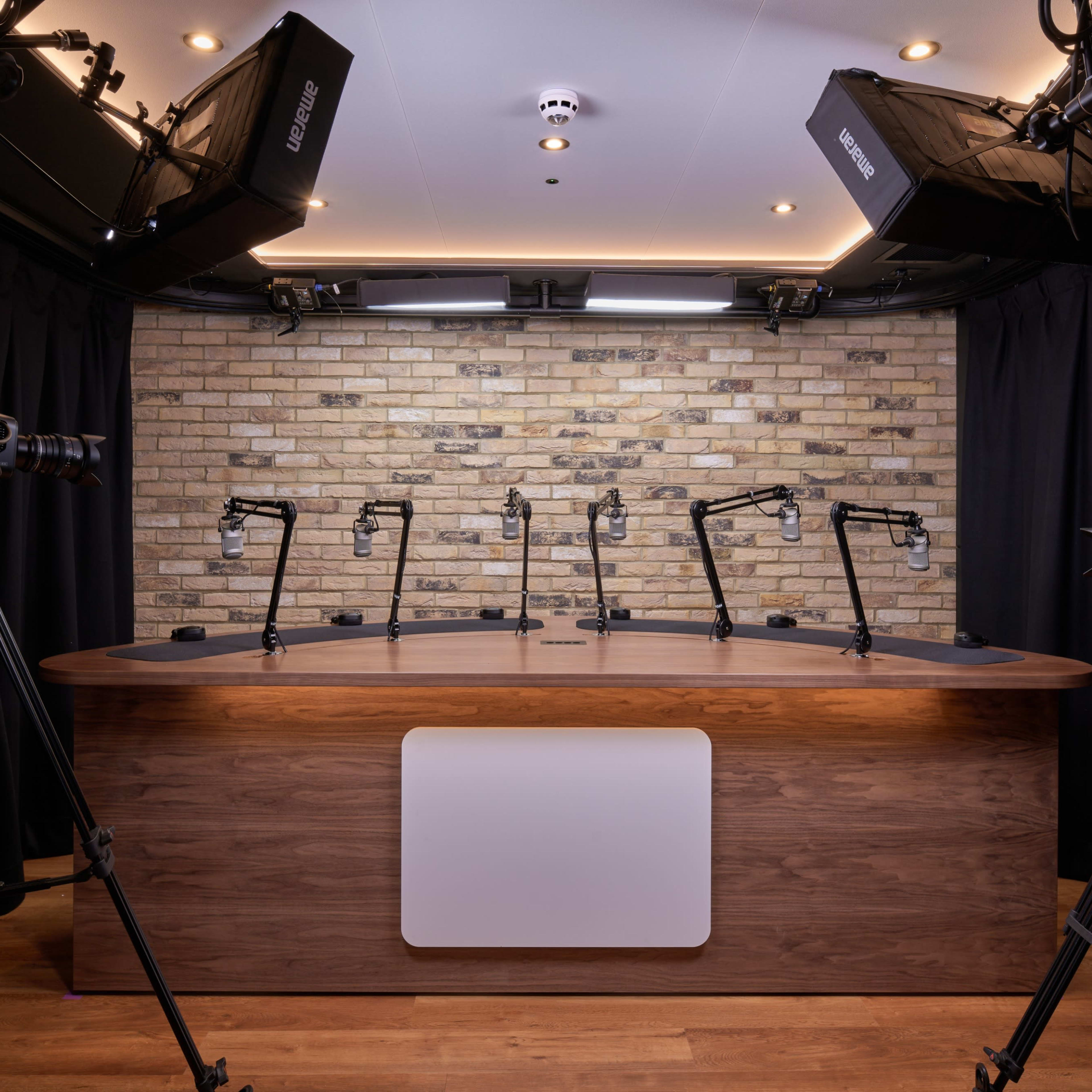 A recording studio featuring a wooden table with multiple microphone stands, surrounded by tripods and softbox lights against a stone wall.
