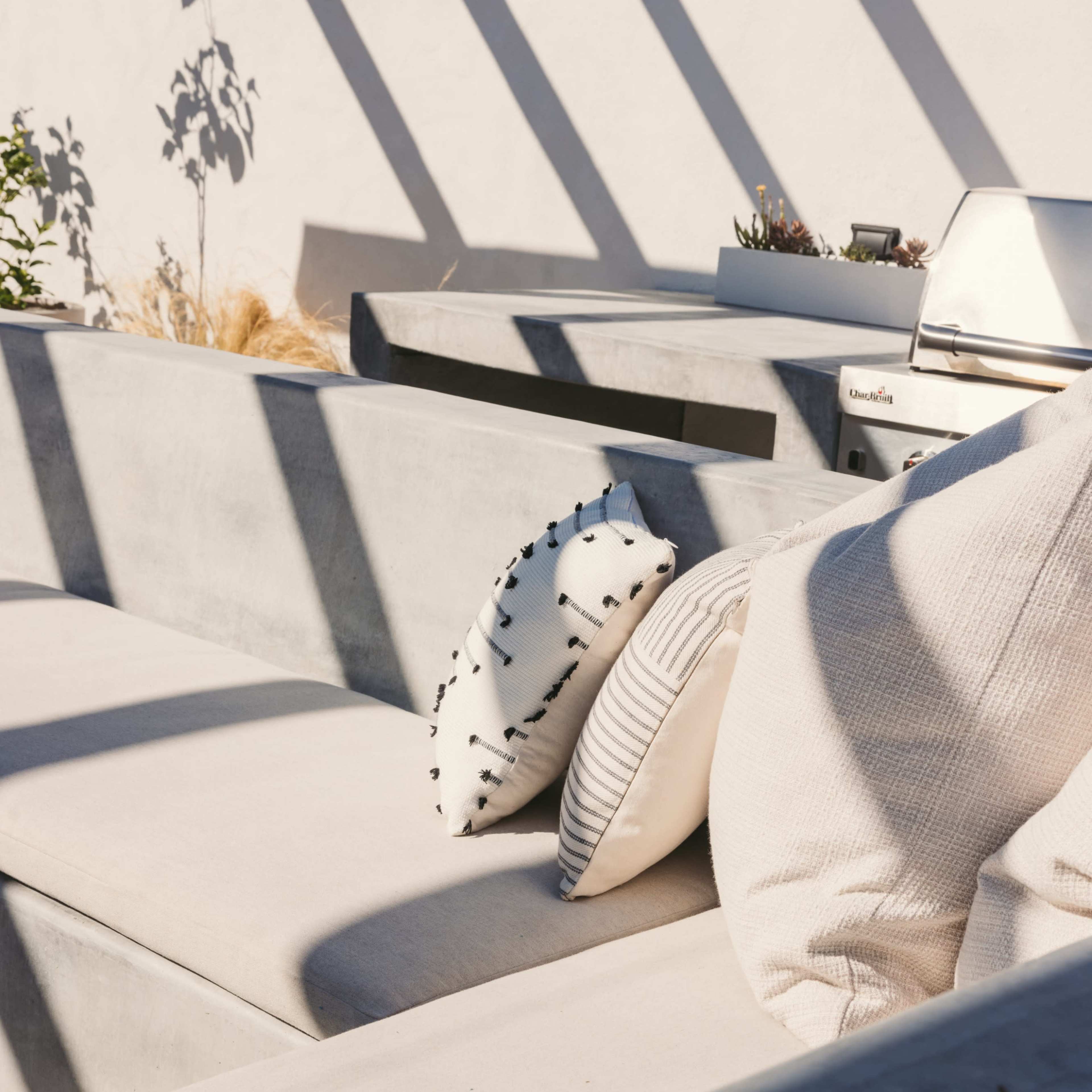 A modern outdoor seating area with concrete benches and decorative cushions, partially shaded by diagonal shadows.