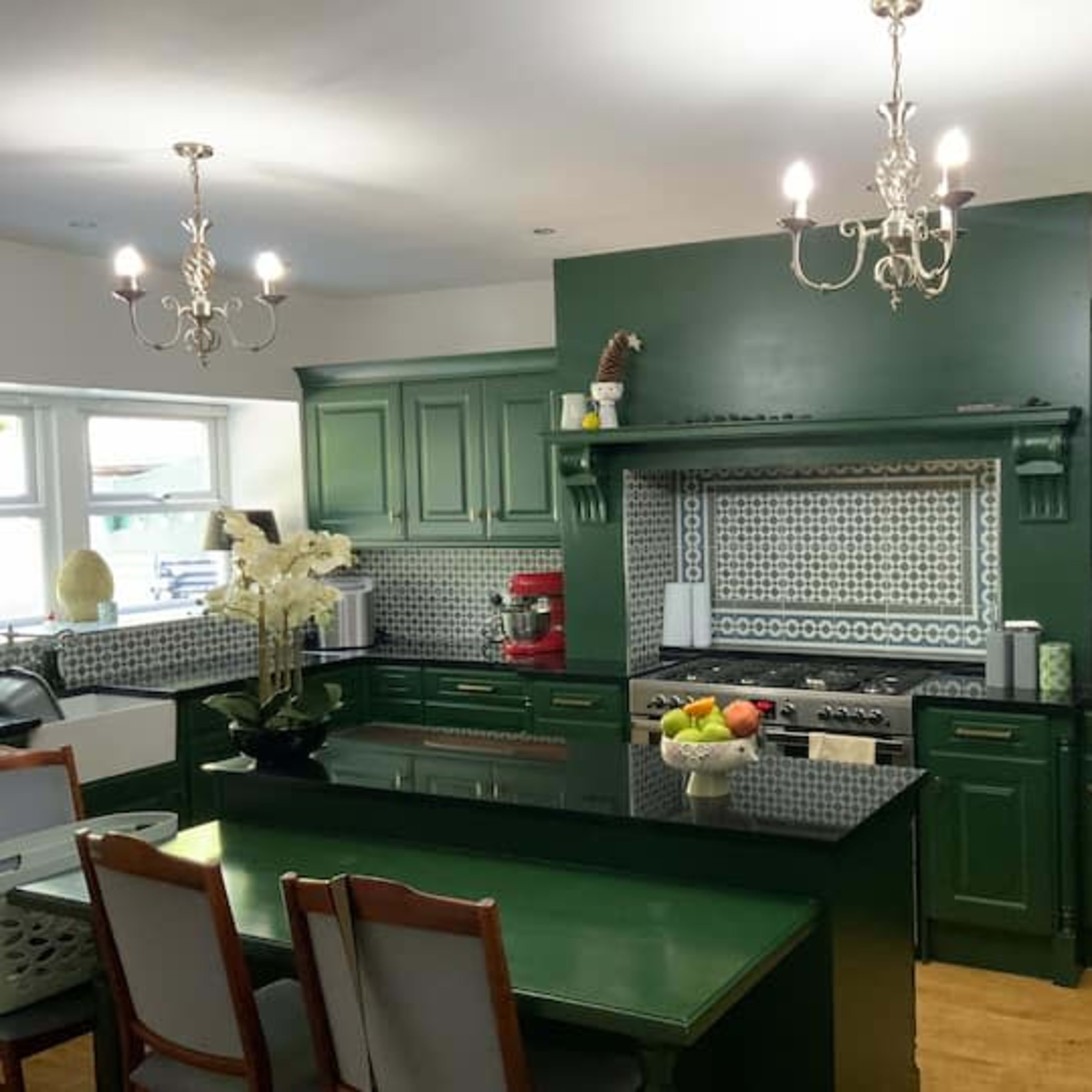The kitchen features green cabinetry, a black countertop, and decorative tile backsplashes with two chandeliers overhead.