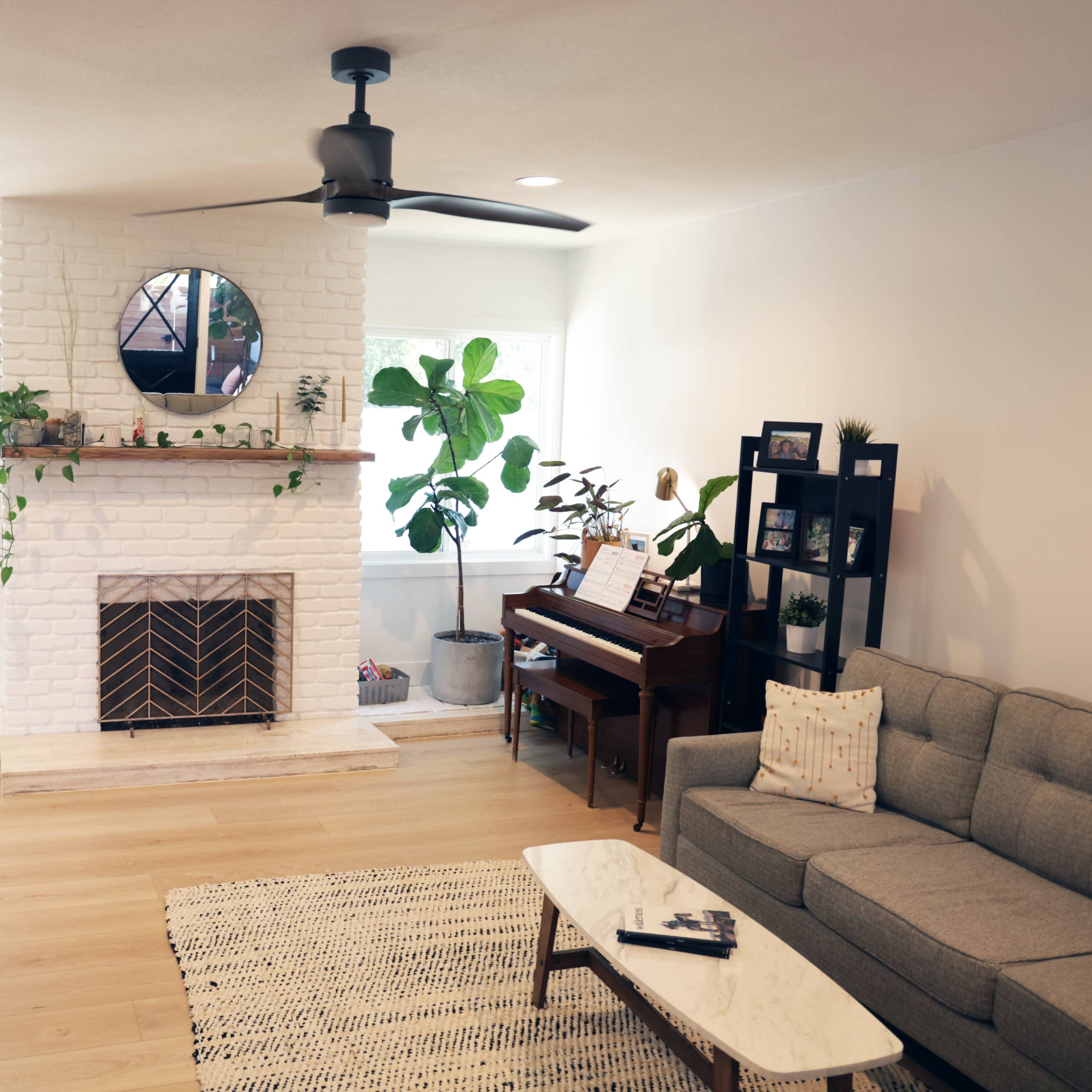 A living room with a gray sofa, a piano, and a white brick fireplace, adorned with plants and a round mirror above the mantel.