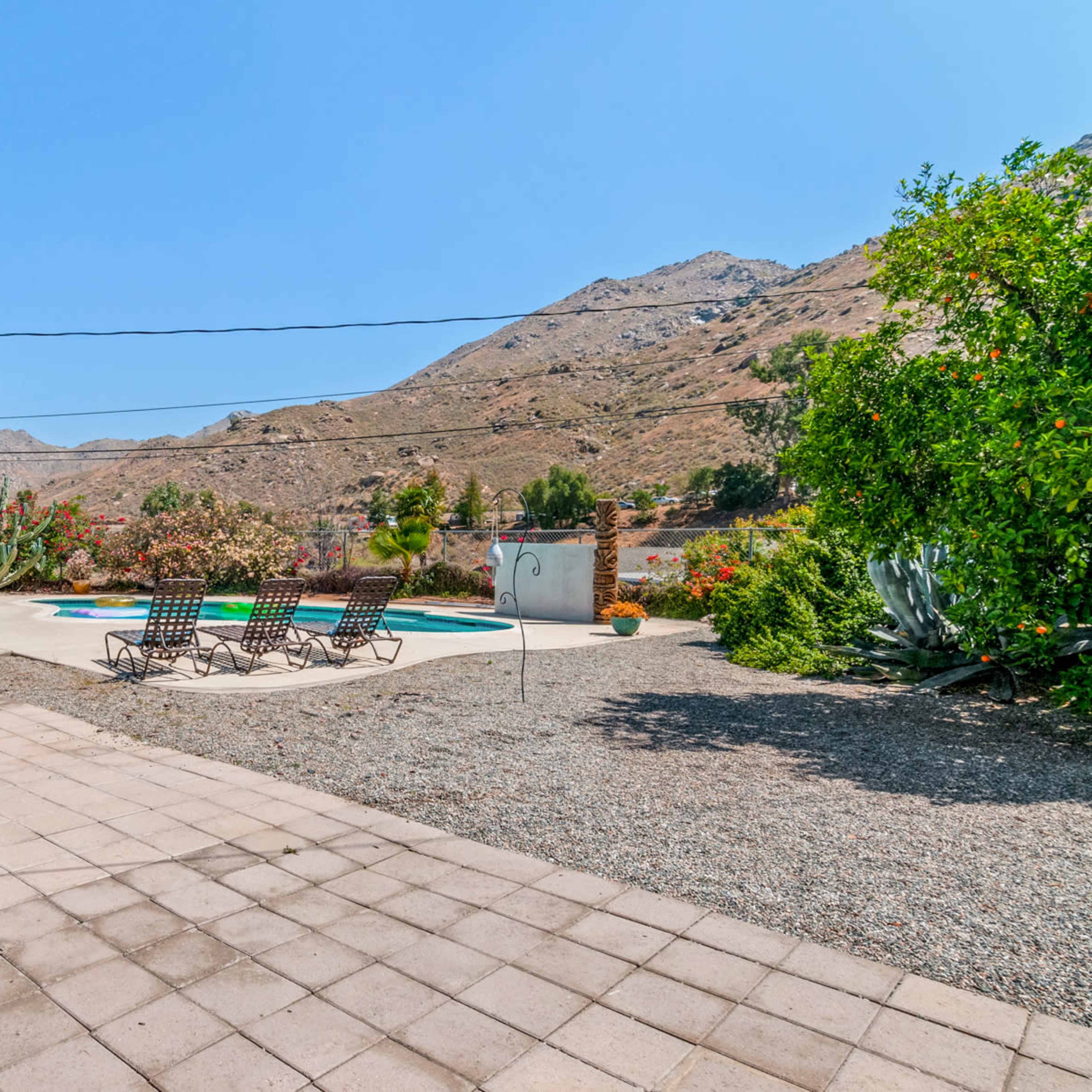 A swimming pool surrounded by lounge chairs is set against a mountainous backdrop with a patio area featuring a table and umbrella.