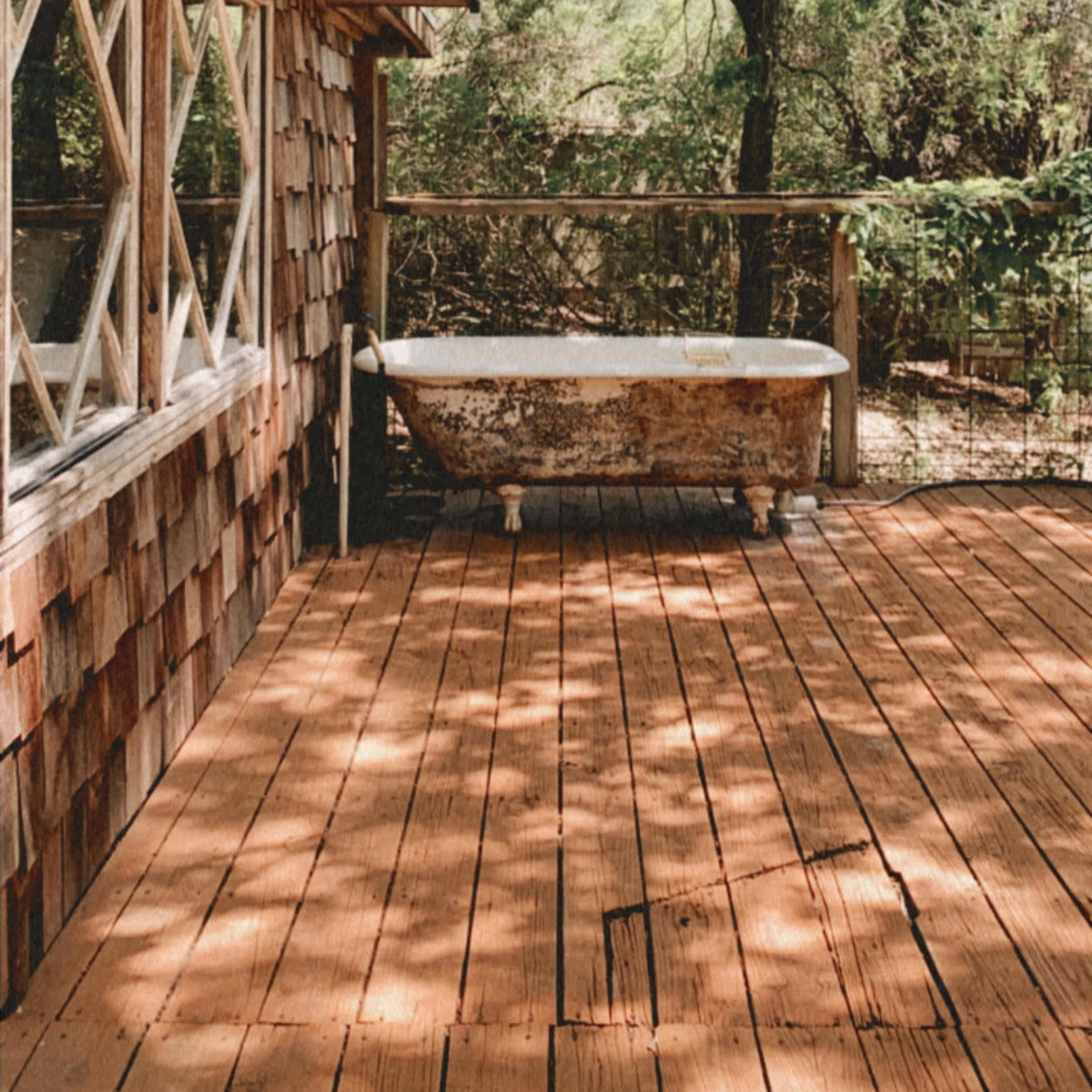 A weathered wooden deck features a vintage bathtub and a backdrop of greenery.