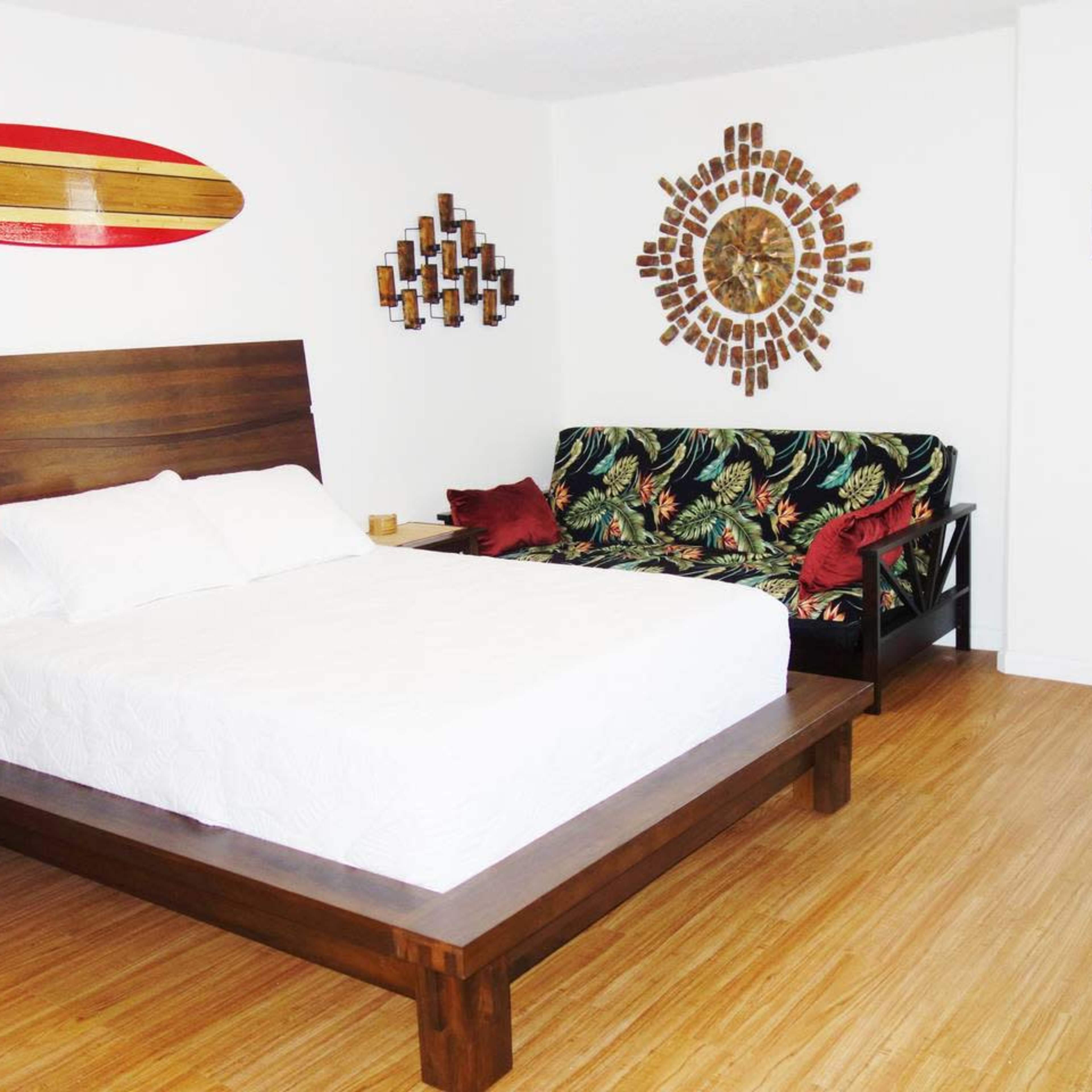 A tidy bedroom featuring a wooden bed, a patterned loveseat, a decorative wall clock, and a surfboard mounted on the wall.