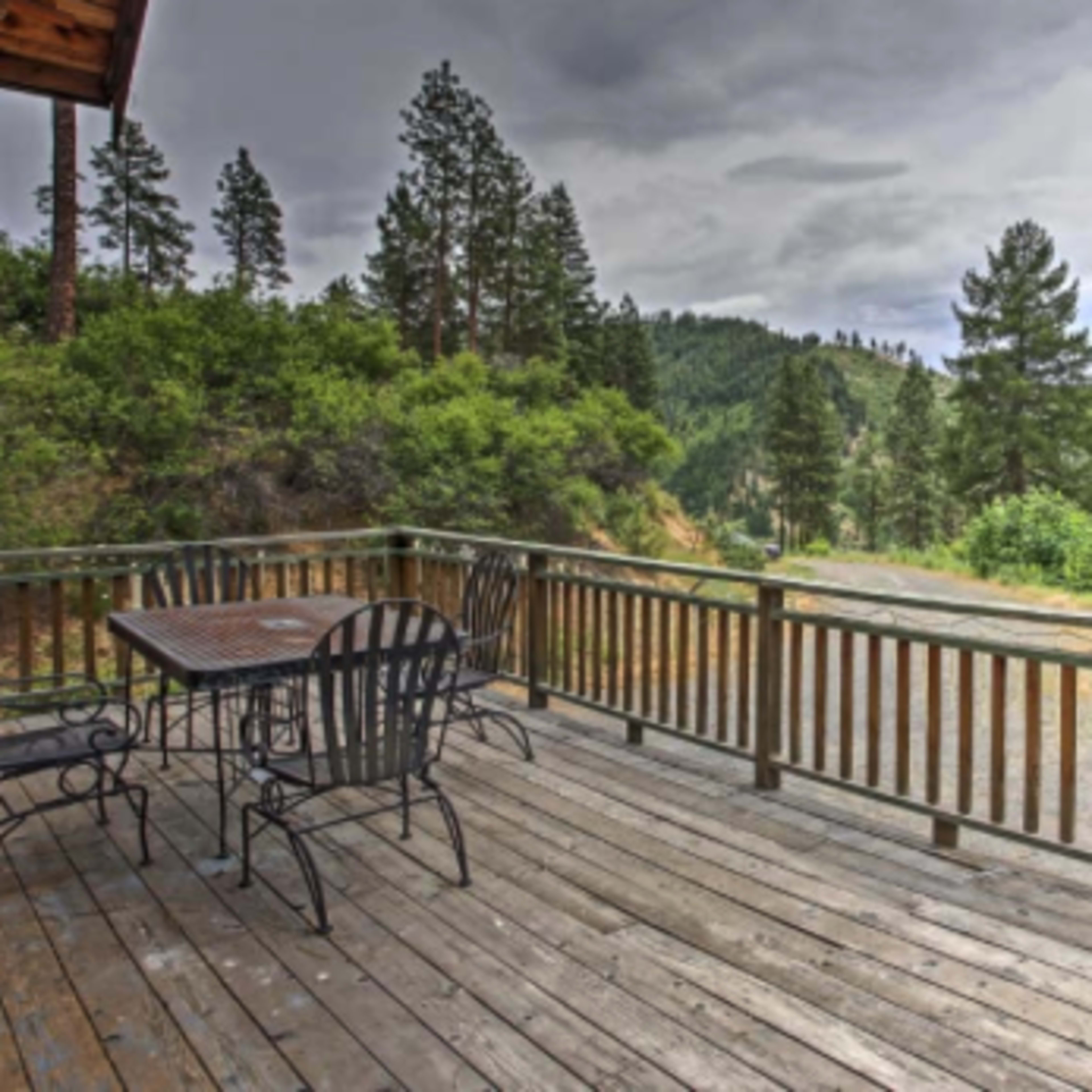 A wooden deck with a metal table and chairs overlooks a mountainous landscape filled with pine trees under a cloudy sky.