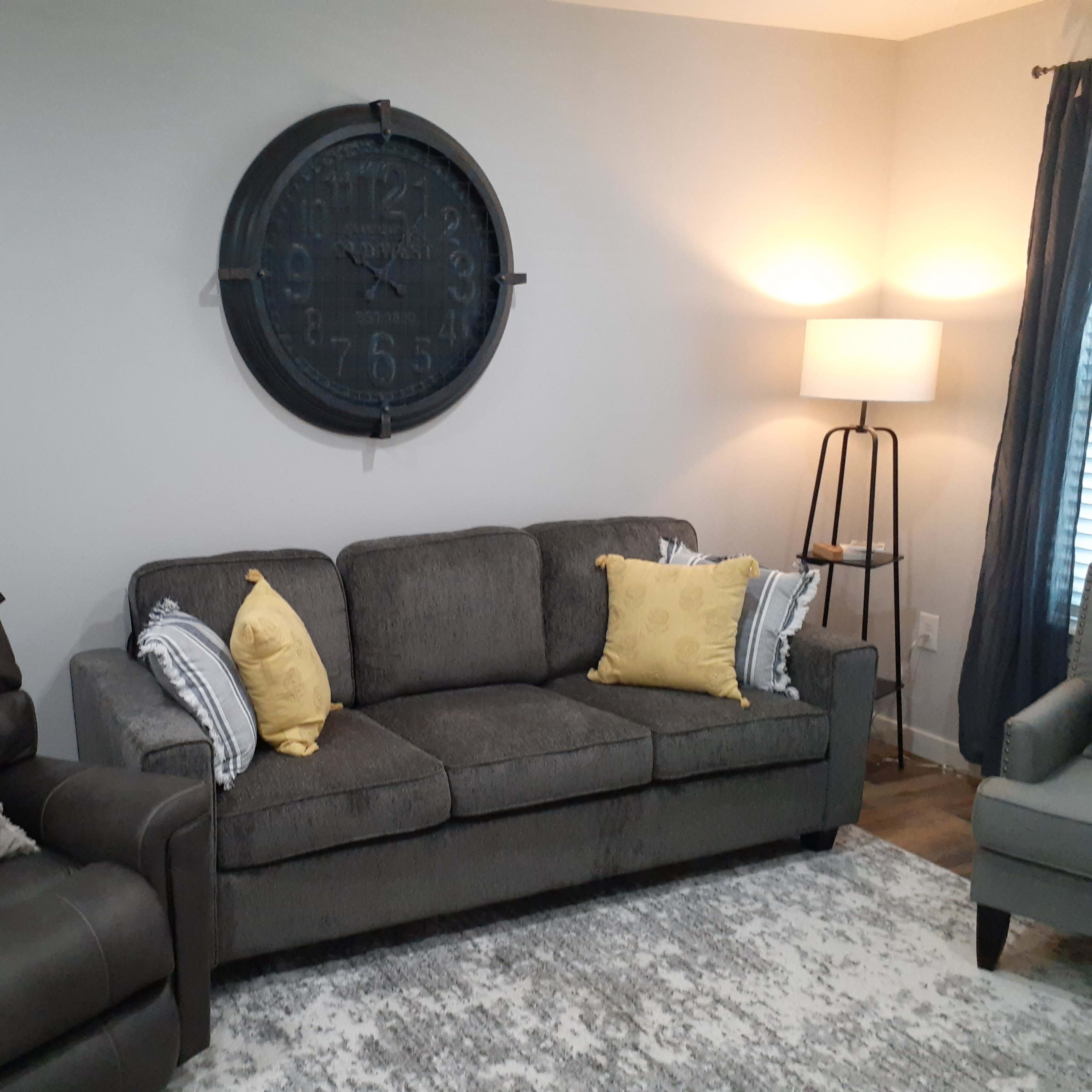 A living room with a gray couch and armchair, a round wall clock, a standing lamp, and yellow decorative pillows on the couch.