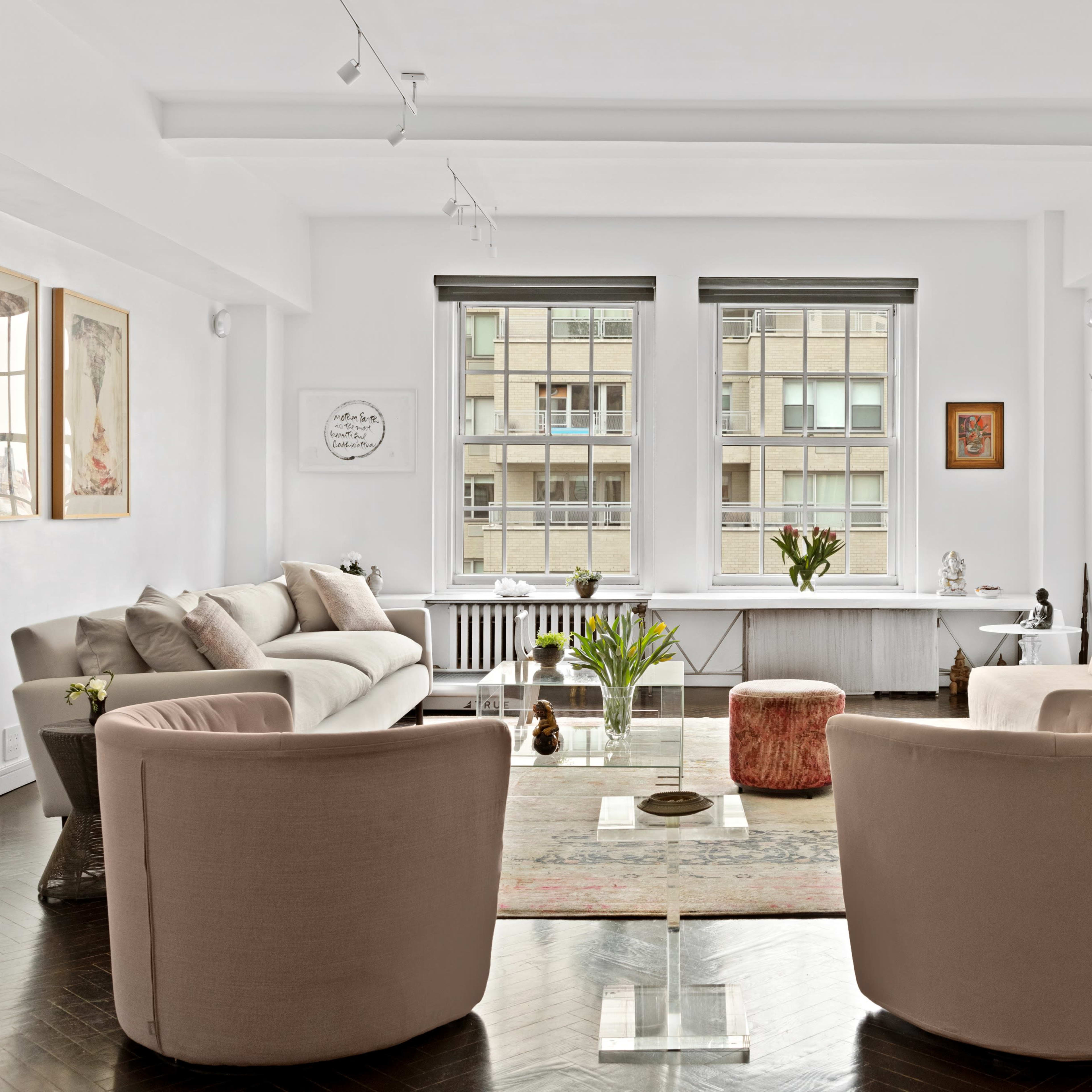 A modern living room features a beige sectional sofa, two armchairs, a glass coffee table, and large windows overlooking an urban view.