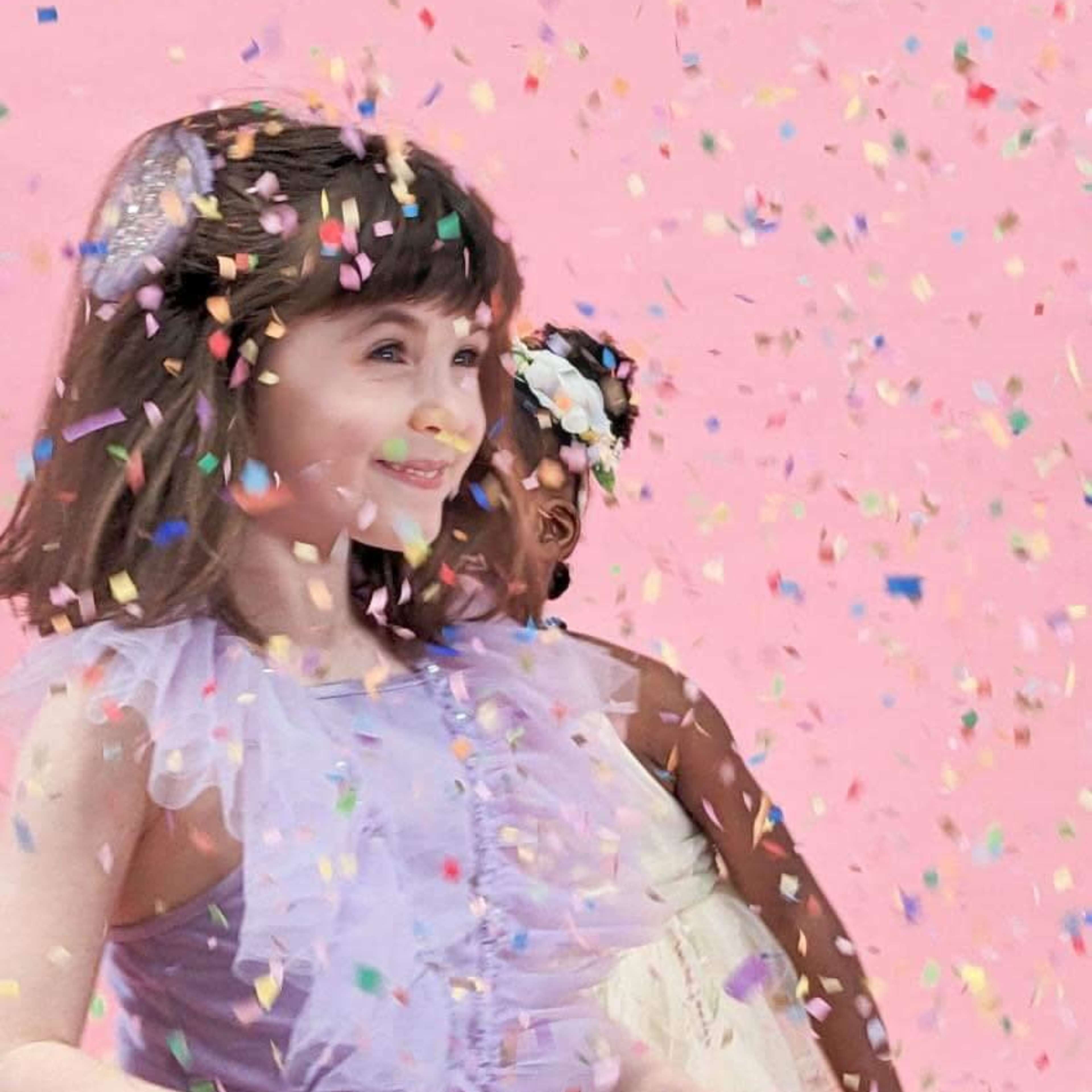 Two young girls smile as colorful confetti falls around them against a pink background.