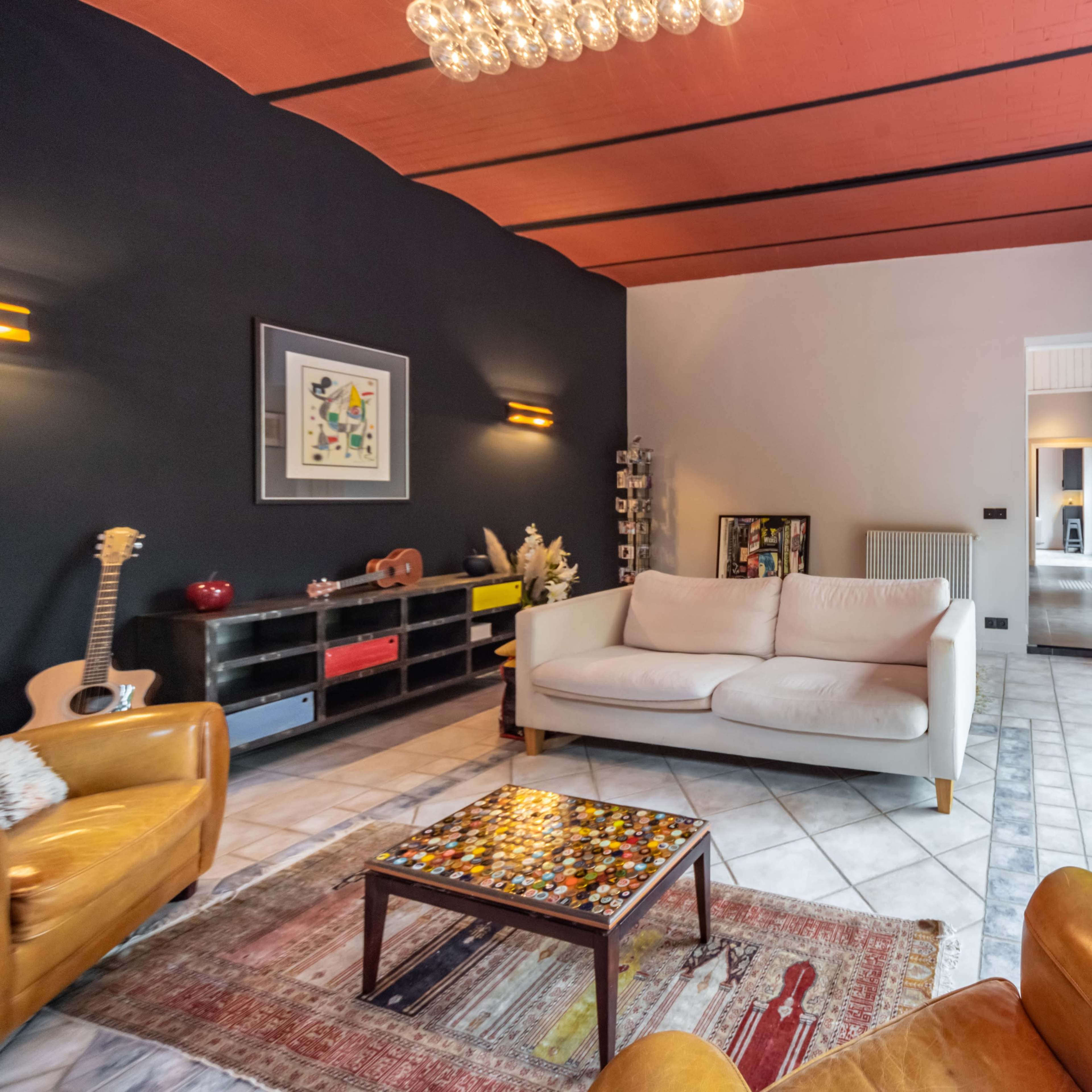 A modern living room features a white sofa, two leather chairs, a colorful coffee table, and large windows overlooking a garden.
