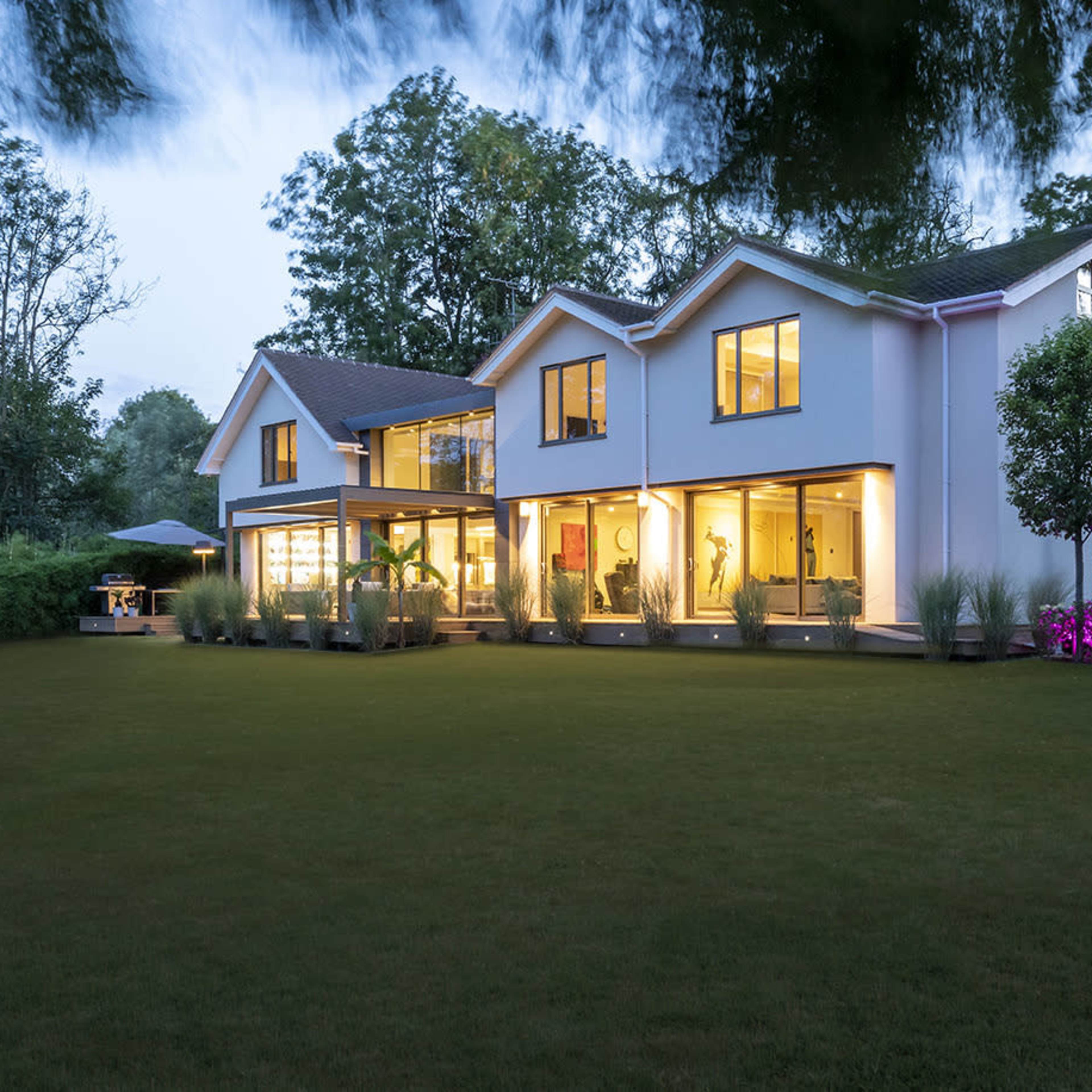 A modern two-story house with large windows is surrounded by a well-maintained lawn and gardens in the evening light.