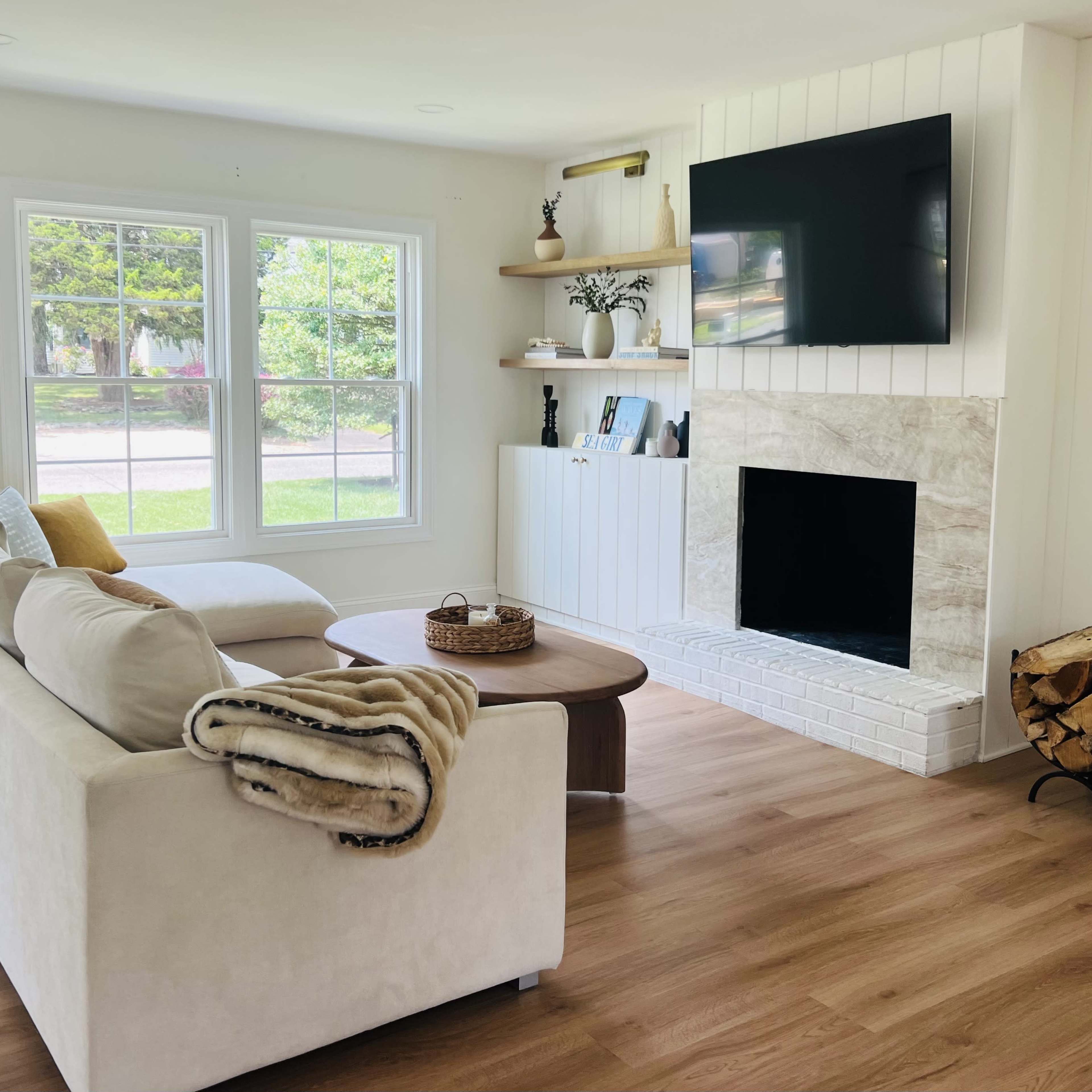 A bright and modern living room featuring a light-colored sofa, a round coffee table, a wall-mounted TV, and a fireplace with stacked firewood beside it.
