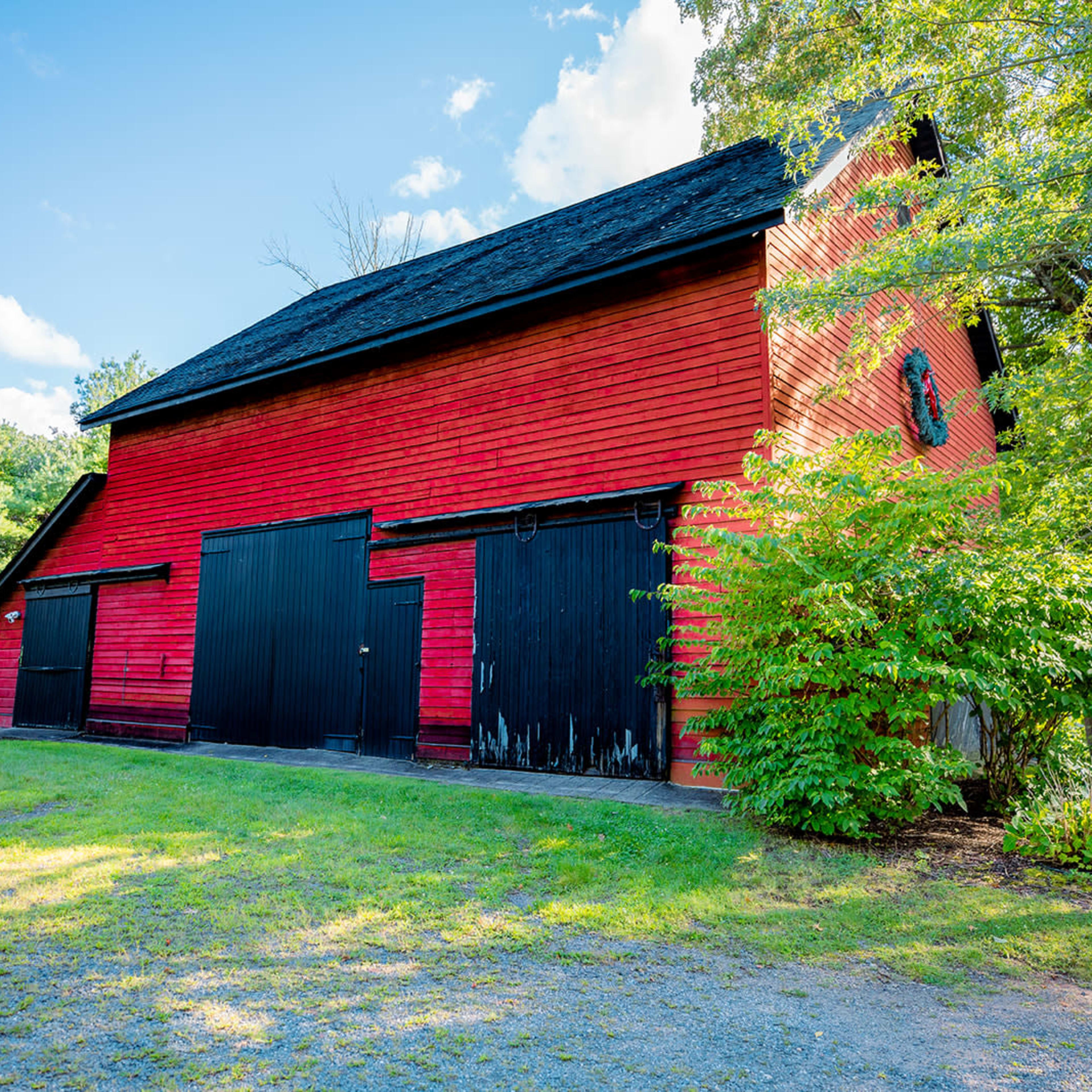 A red barn with black doors stands next to a patch of green grass and trees against a clear blue sky.