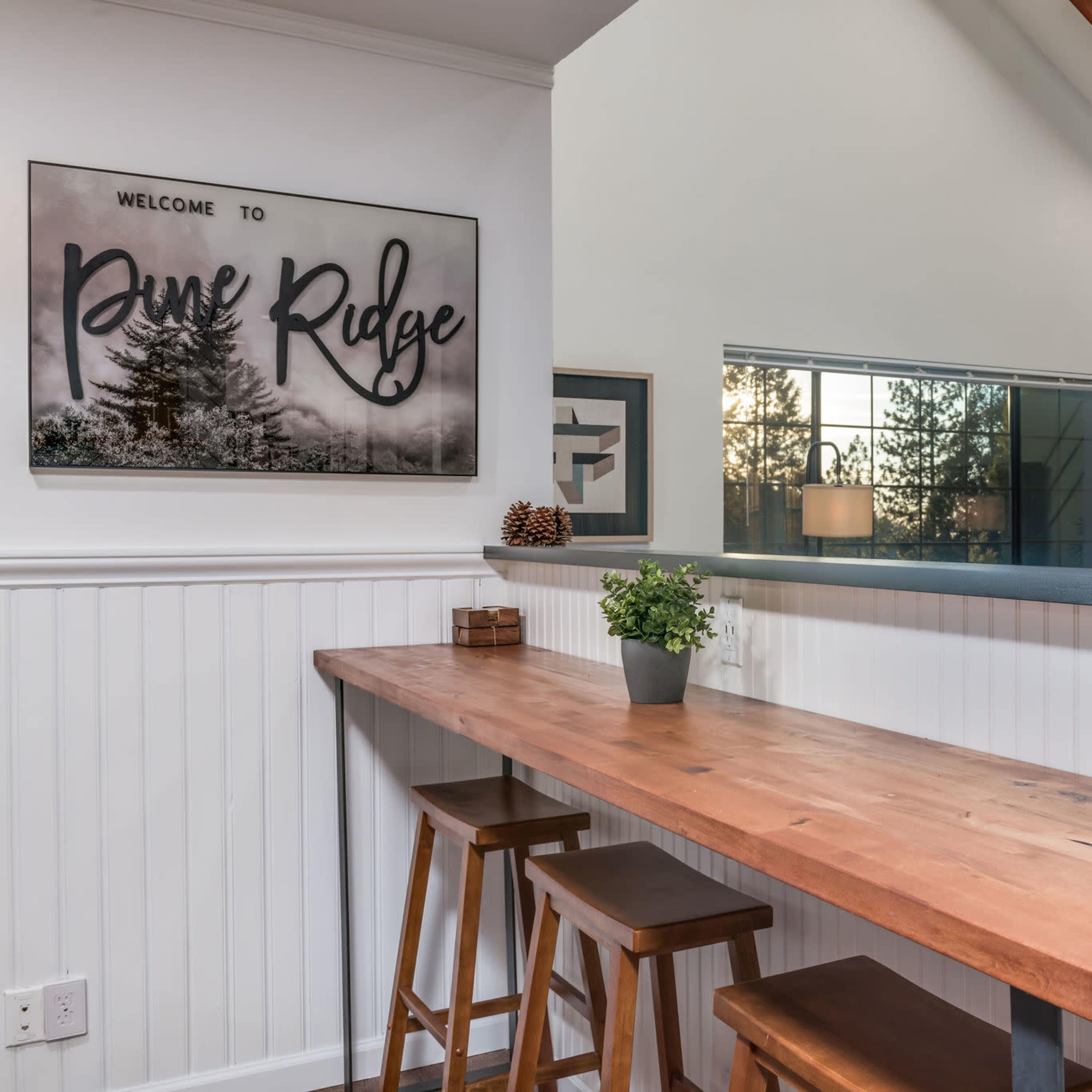 A wooden bar table with three stools sits against a white paneled wall, featuring a wall art piece that reads "Welcome to Pine Ridge."