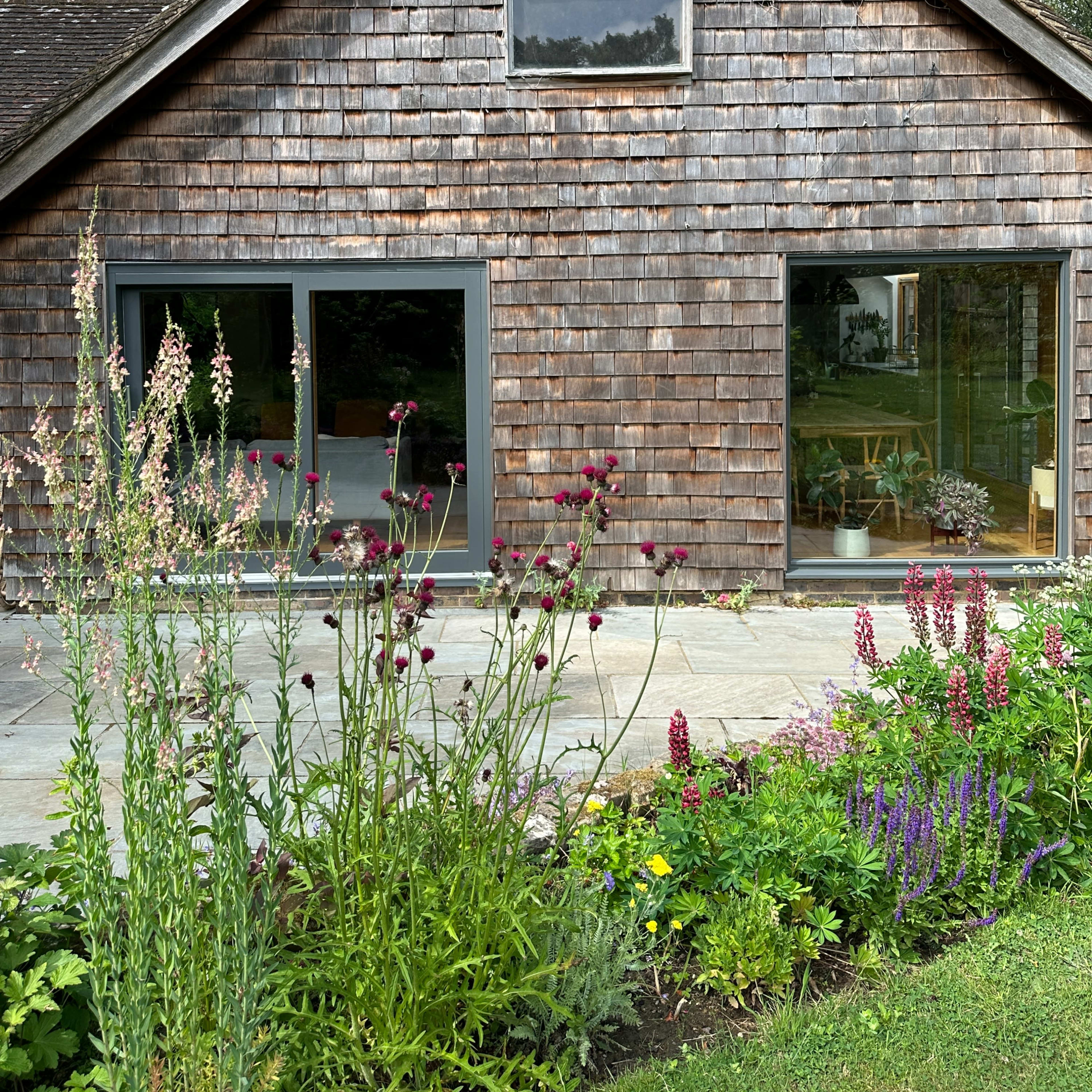 A wooden house with a shingled exterior features large windows, surrounded by a well-maintained garden with various flowering plants.