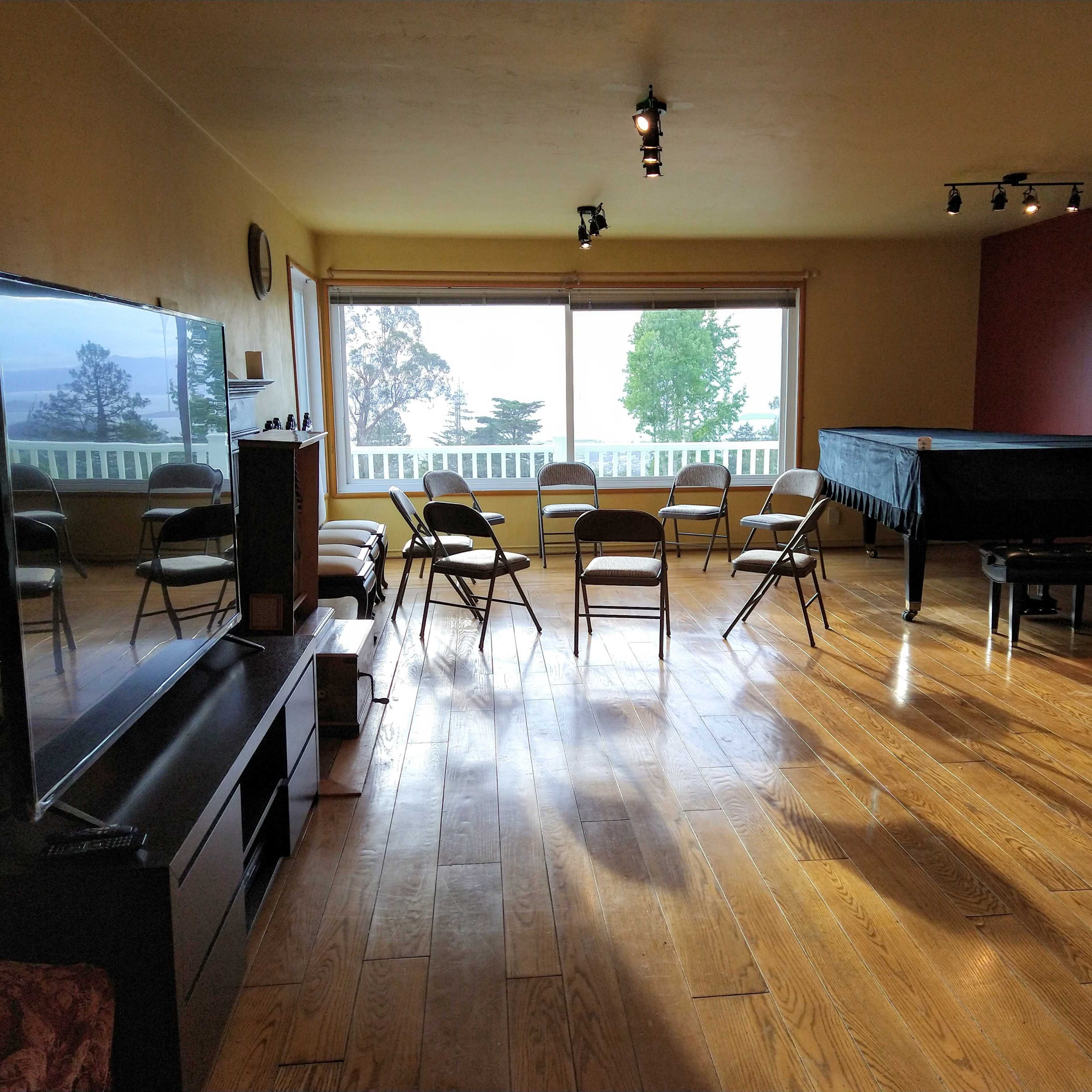 A large, empty room with wooden floors features a set of eight folding chairs arranged in a circle, a grand piano in the corner, and a large window that overlooks a scenic view.