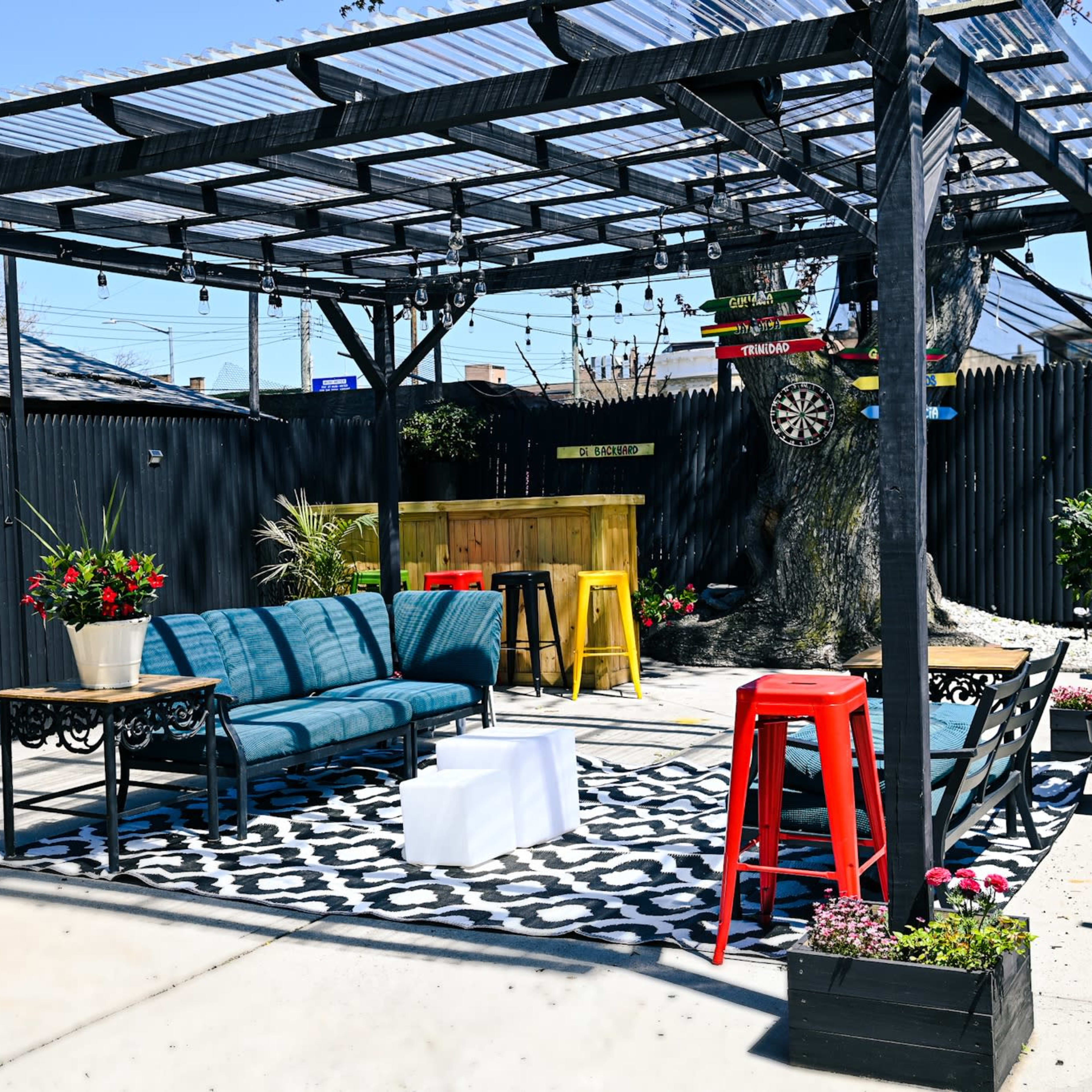 A patio area with a covered seating arrangement, featuring blue sofas, a coffee table, and bright red stools, surrounded by decorative plants and a dartboard.
