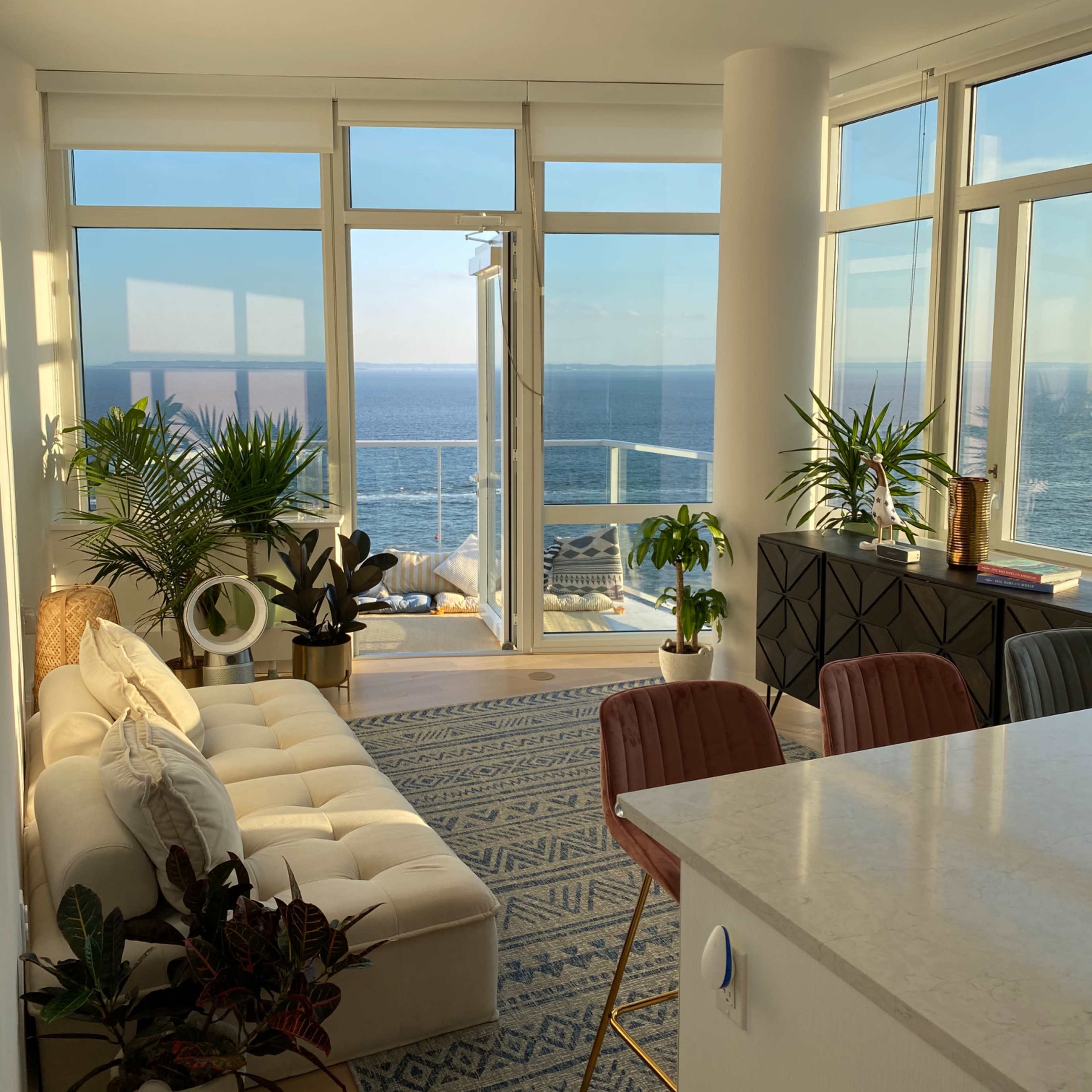 A bright living space features a plush sofa, indoor plants, and a large window overlooking the ocean.