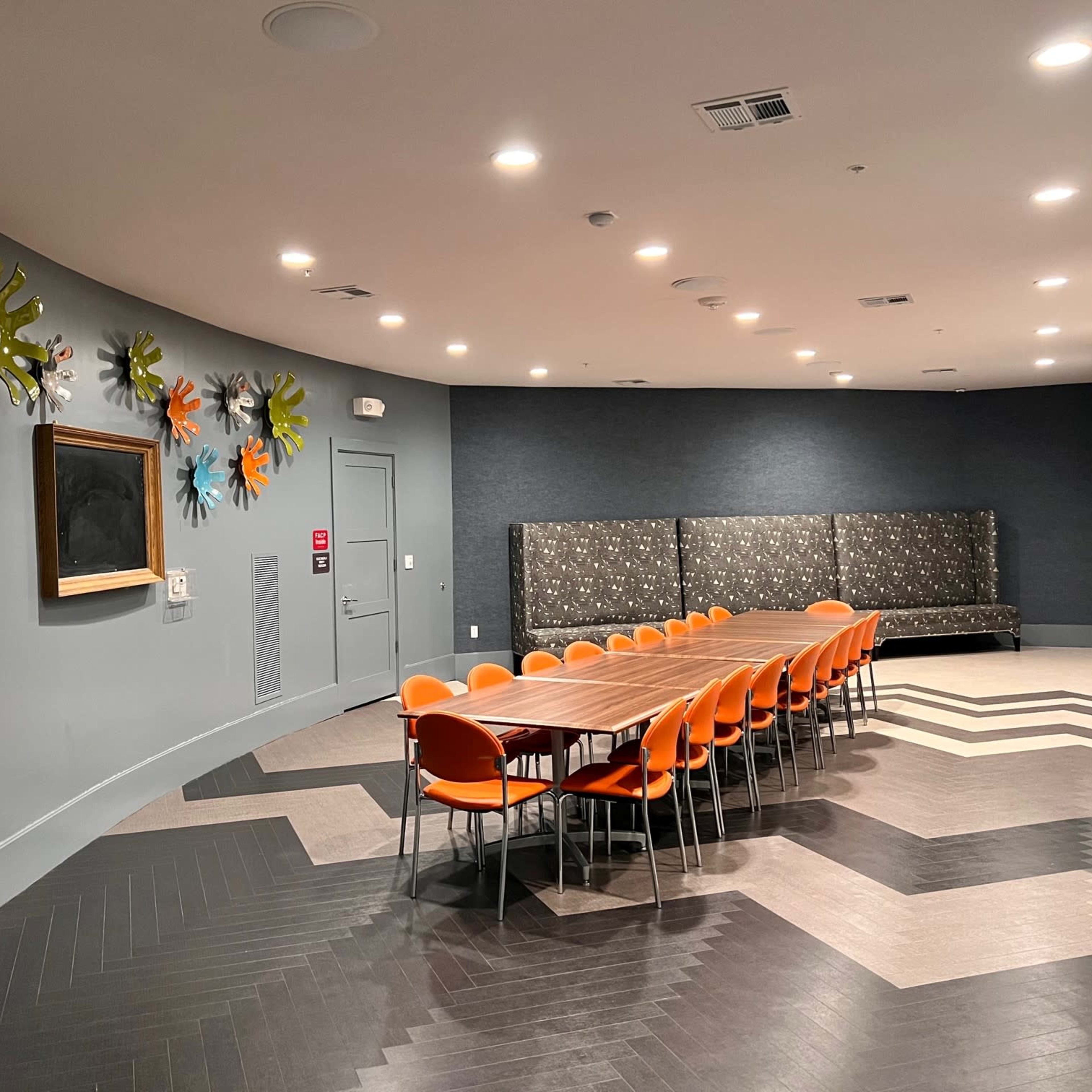 A conference room features a long wooden table surrounded by orange chairs, with colorful wall art and a seating area in the background.