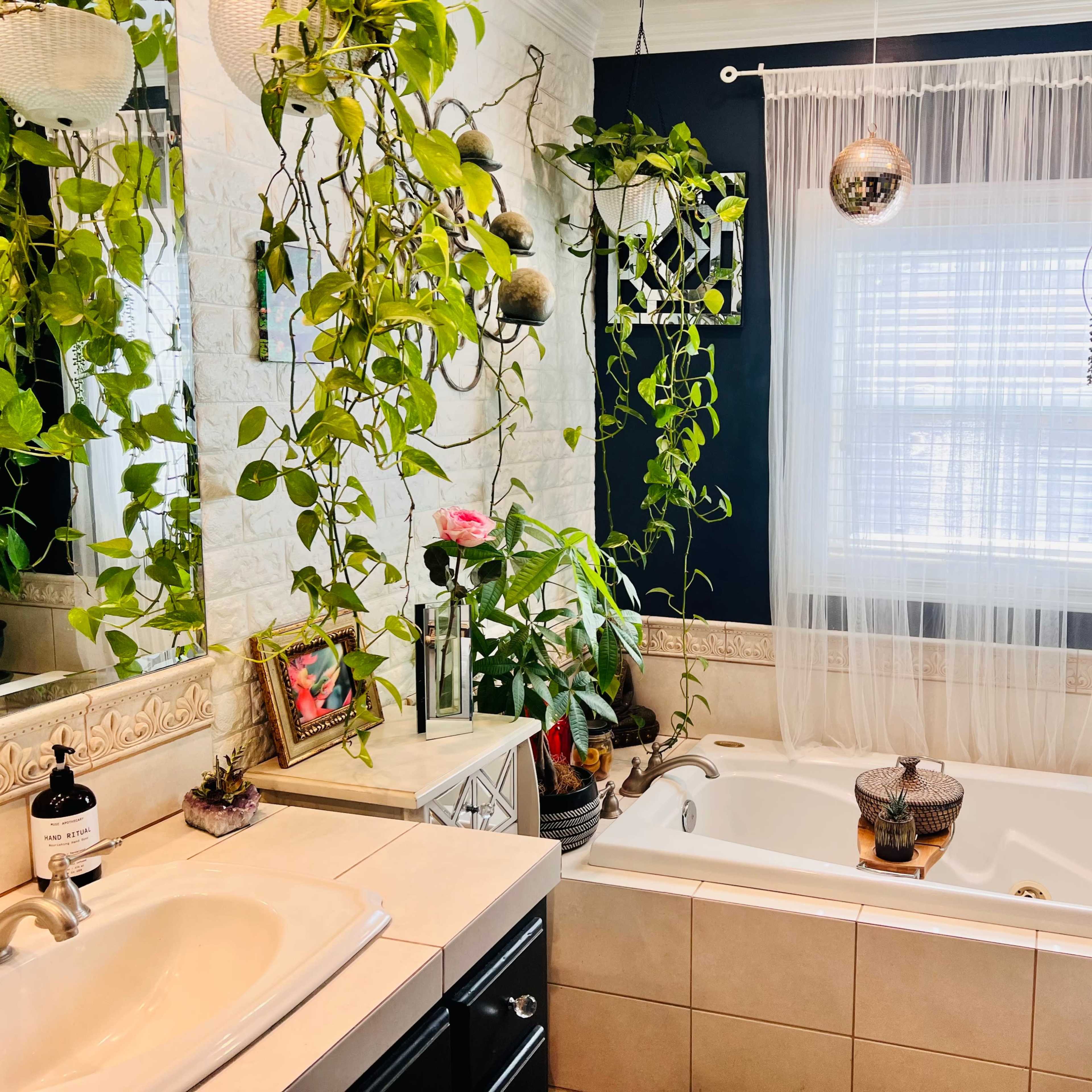 A well-lit bathroom features a white tub surrounded by potted plants and decorative elements near a window dressed with sheer curtains.