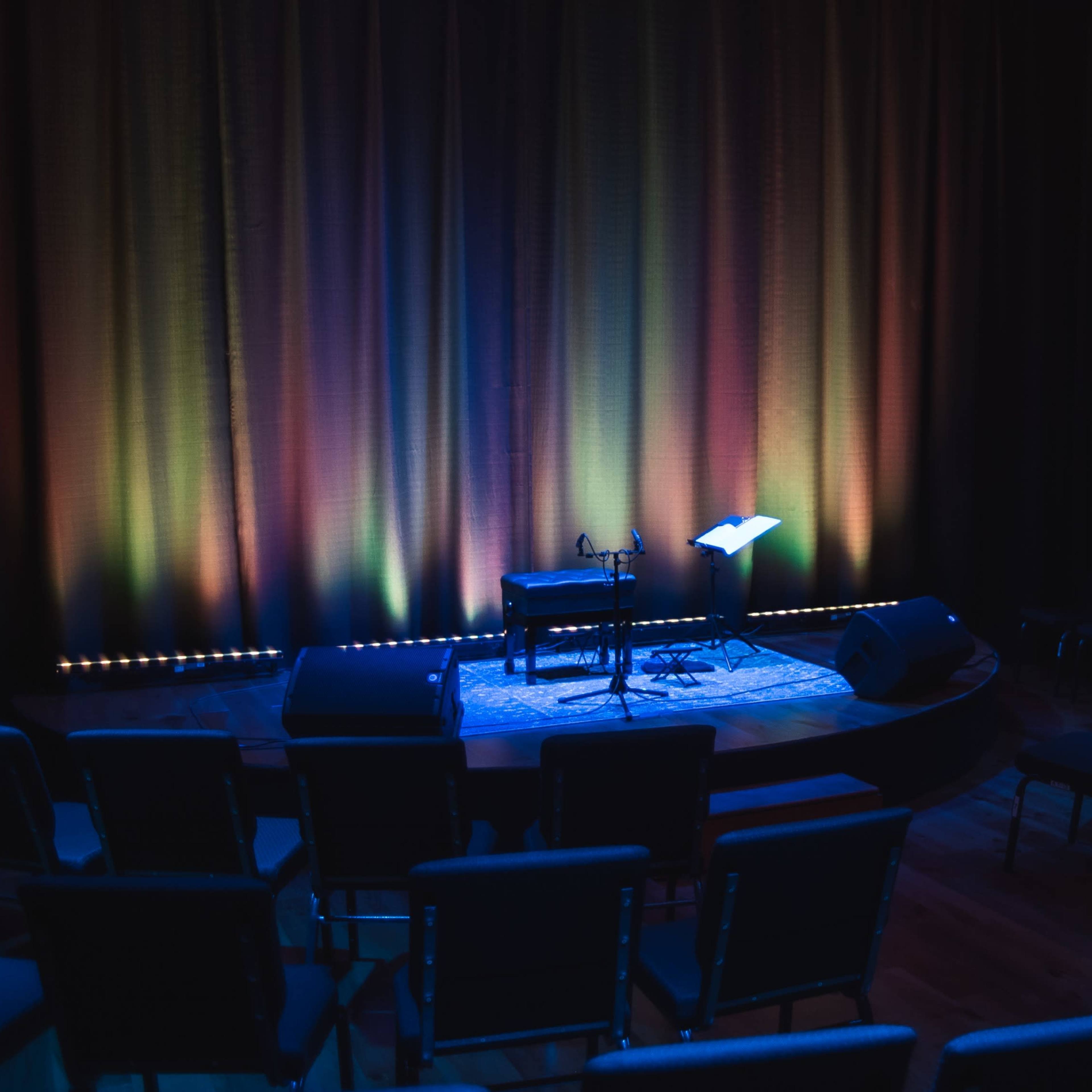 A small stage is set with a microphone and a music stand, surrounded by empty chairs under dim lighting.