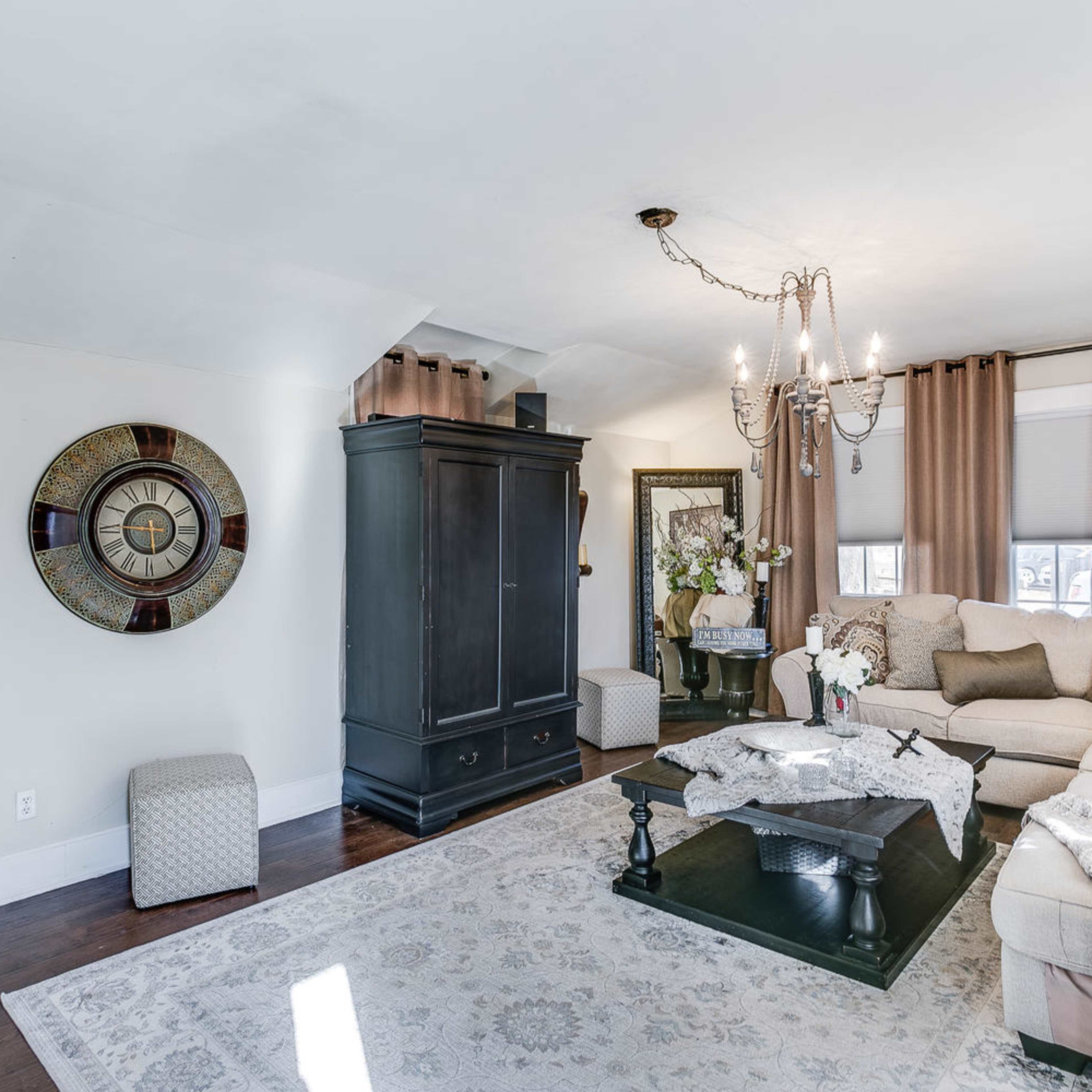 A living room featuring a light-colored sofa, a dark wooden coffee table, a large decorative wall clock, and a chandelier hanging from the ceiling.