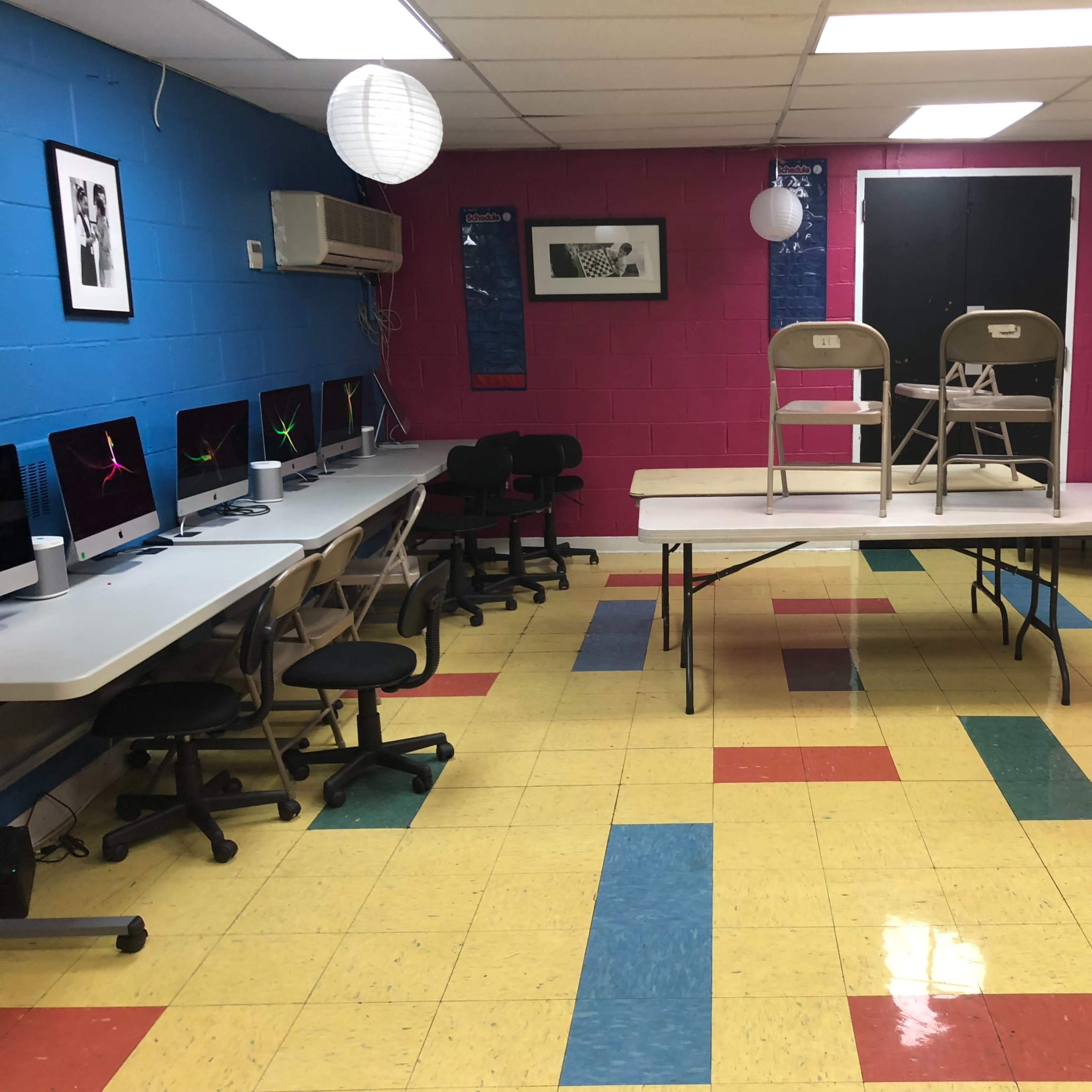 A computer room with rows of desks, chairs, and a table displaying stacked chairs, set against a brightly colored wall.