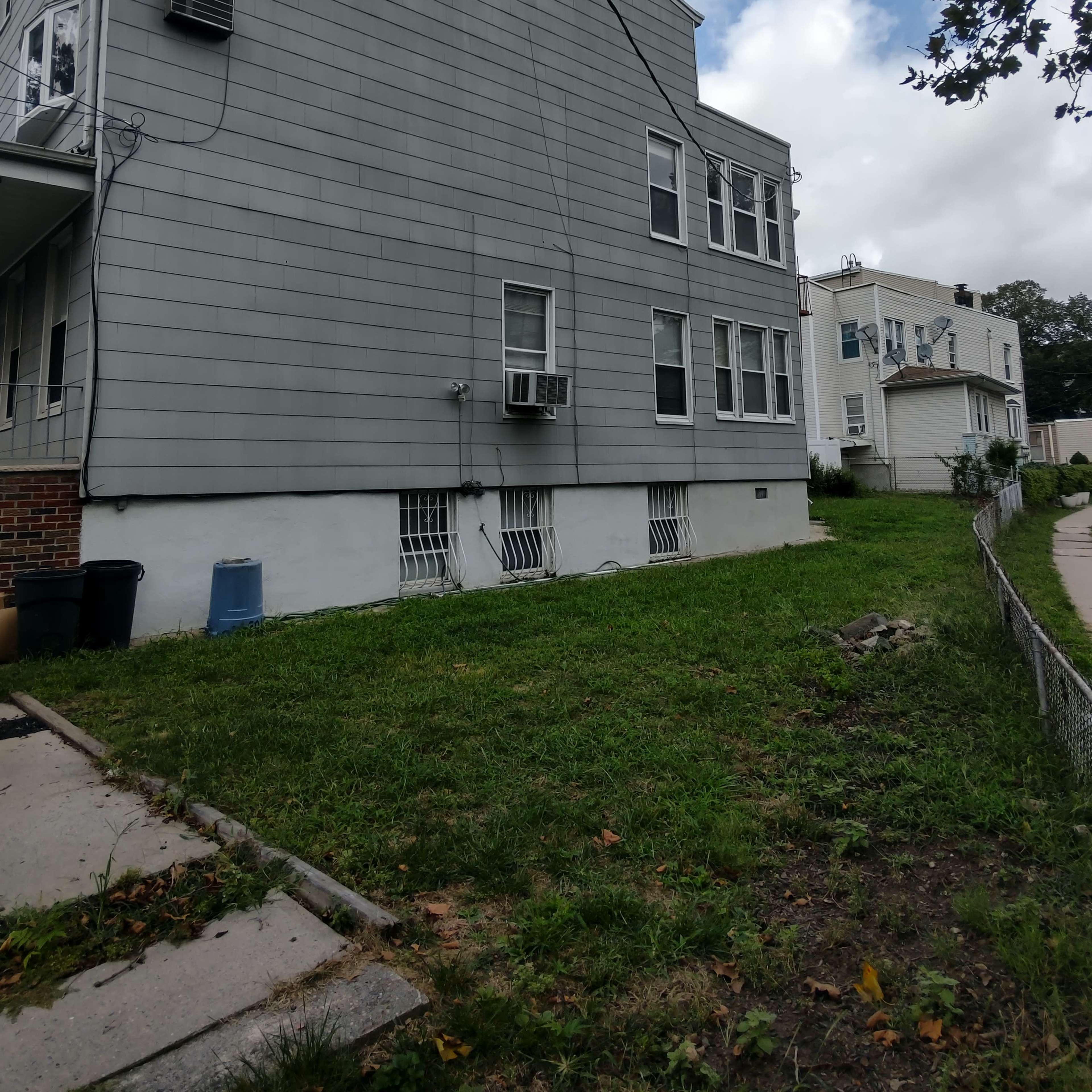 A gray, two-story house with multiple windows and air conditioning units, situated on a grassy yard next to a sidewalk.