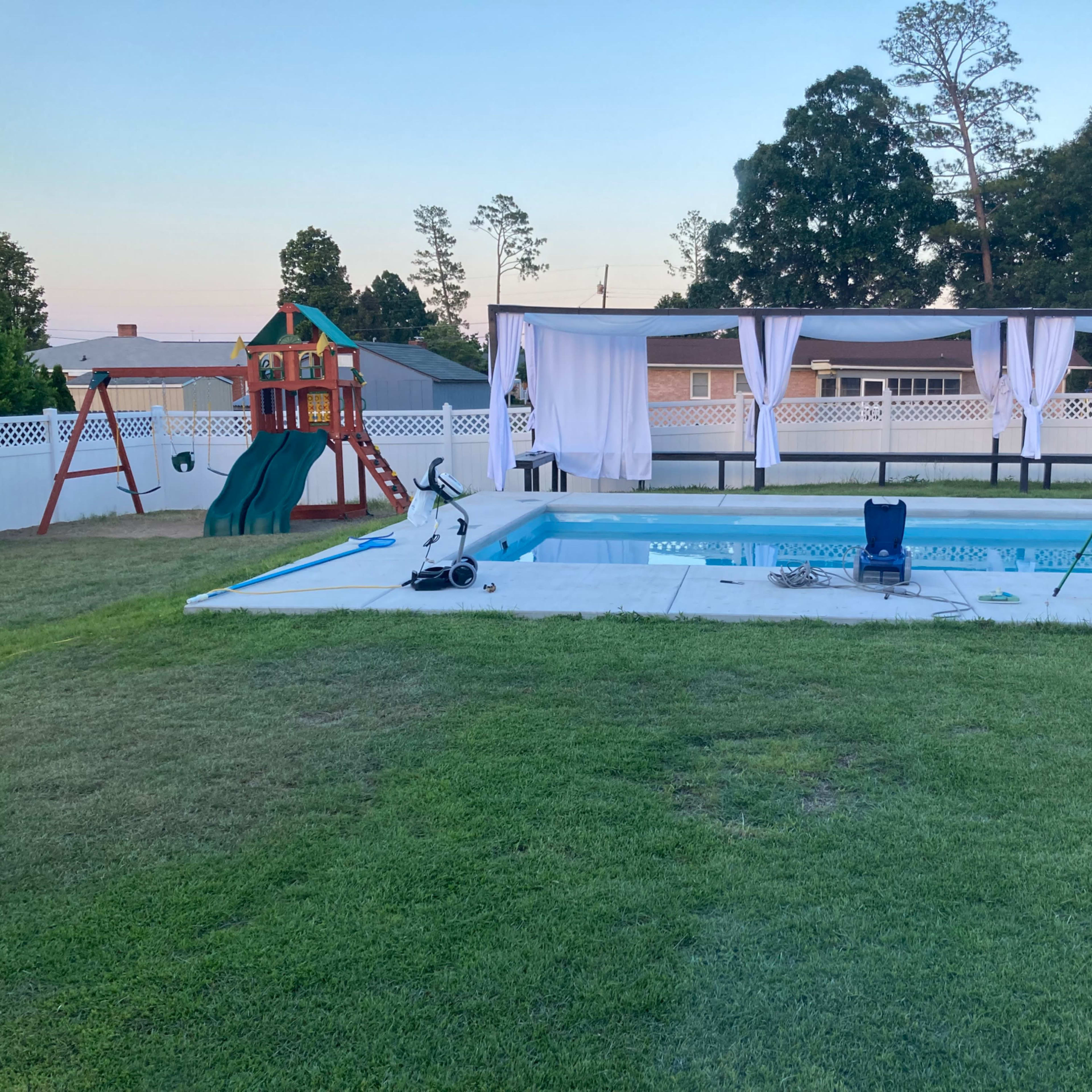 A backyard with a swimming pool, a covered seating area, a playground structure, and green grass.