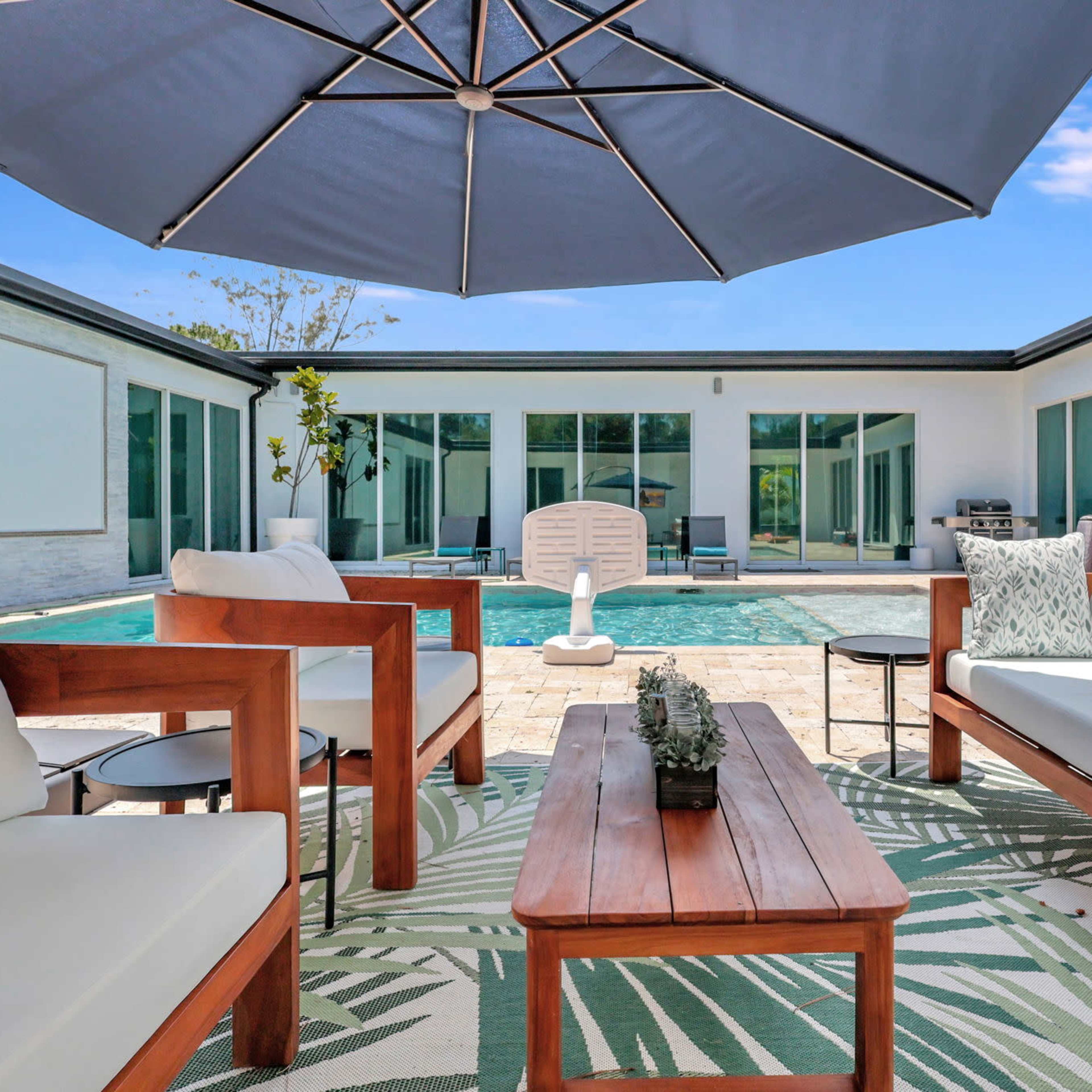 A backyard seating area with wooden furniture and a large umbrella beside a swimming pool surrounded by glass-walled rooms.