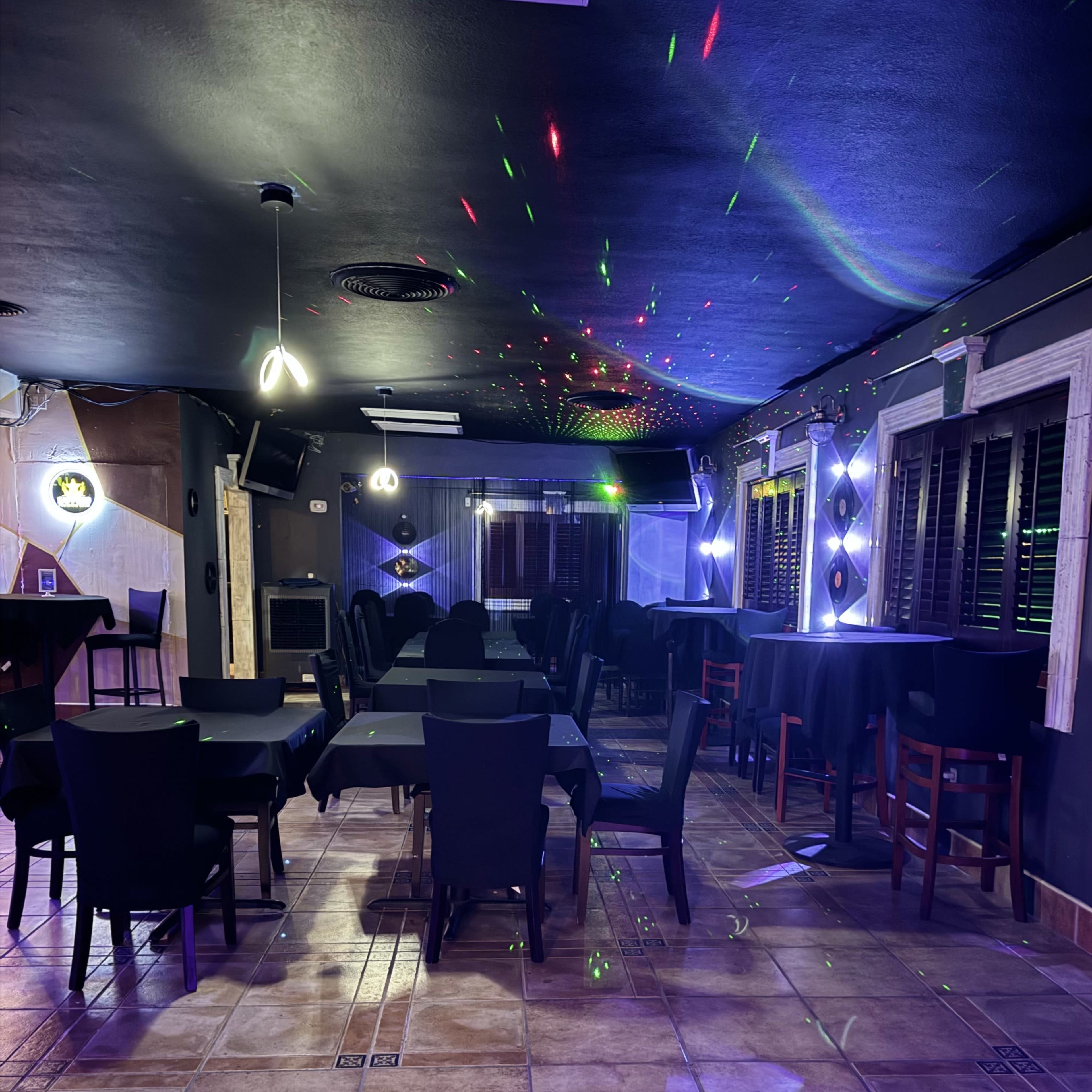 The interior of a dimly lit restaurant features black tables and chairs, with colorful lights reflecting on the walls and ceiling.