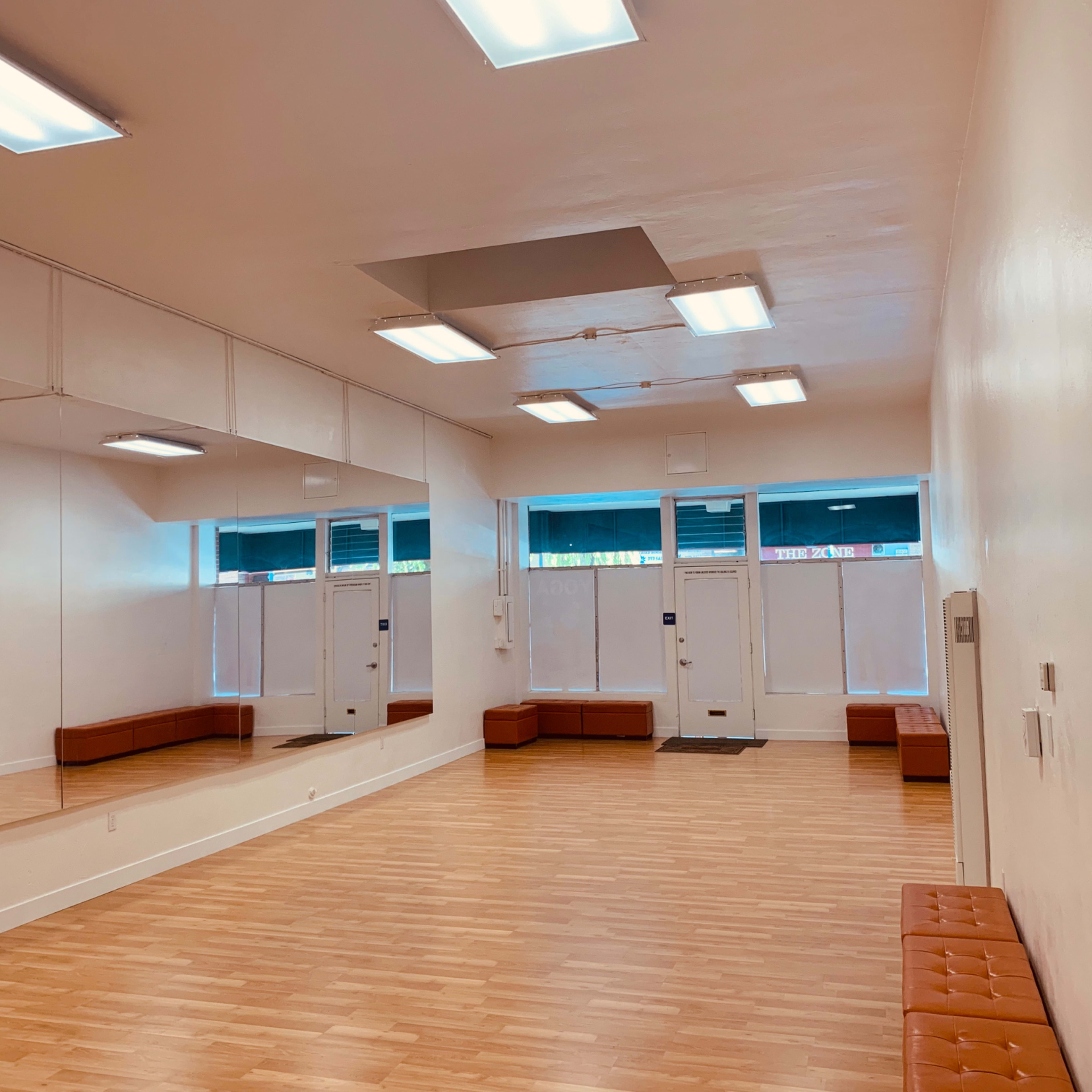 A spacious, empty room with wooden flooring, large mirrors on one wall, and two entrance doors at the front.