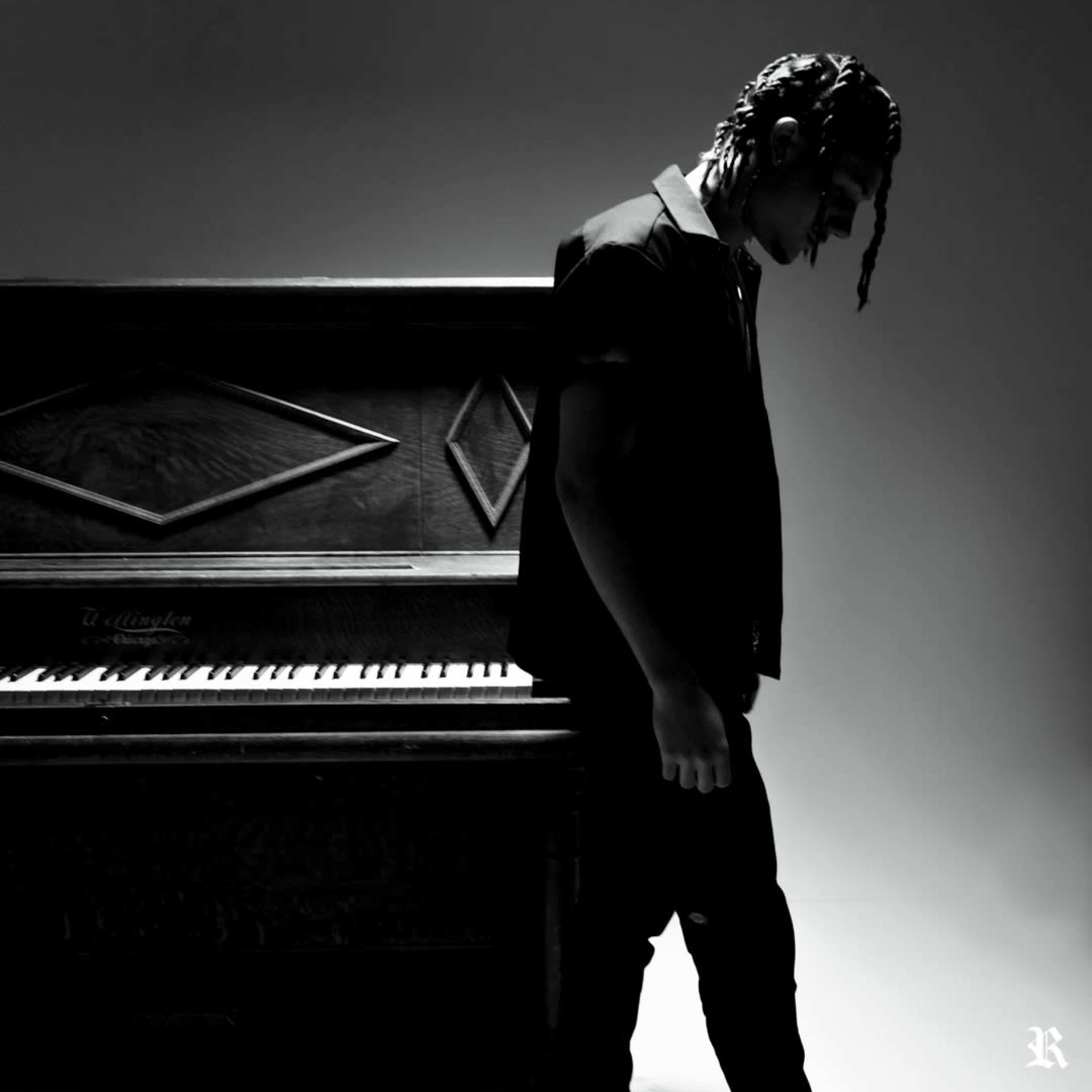 A person stands beside an upright piano in a dimly lit space, gazing downward.