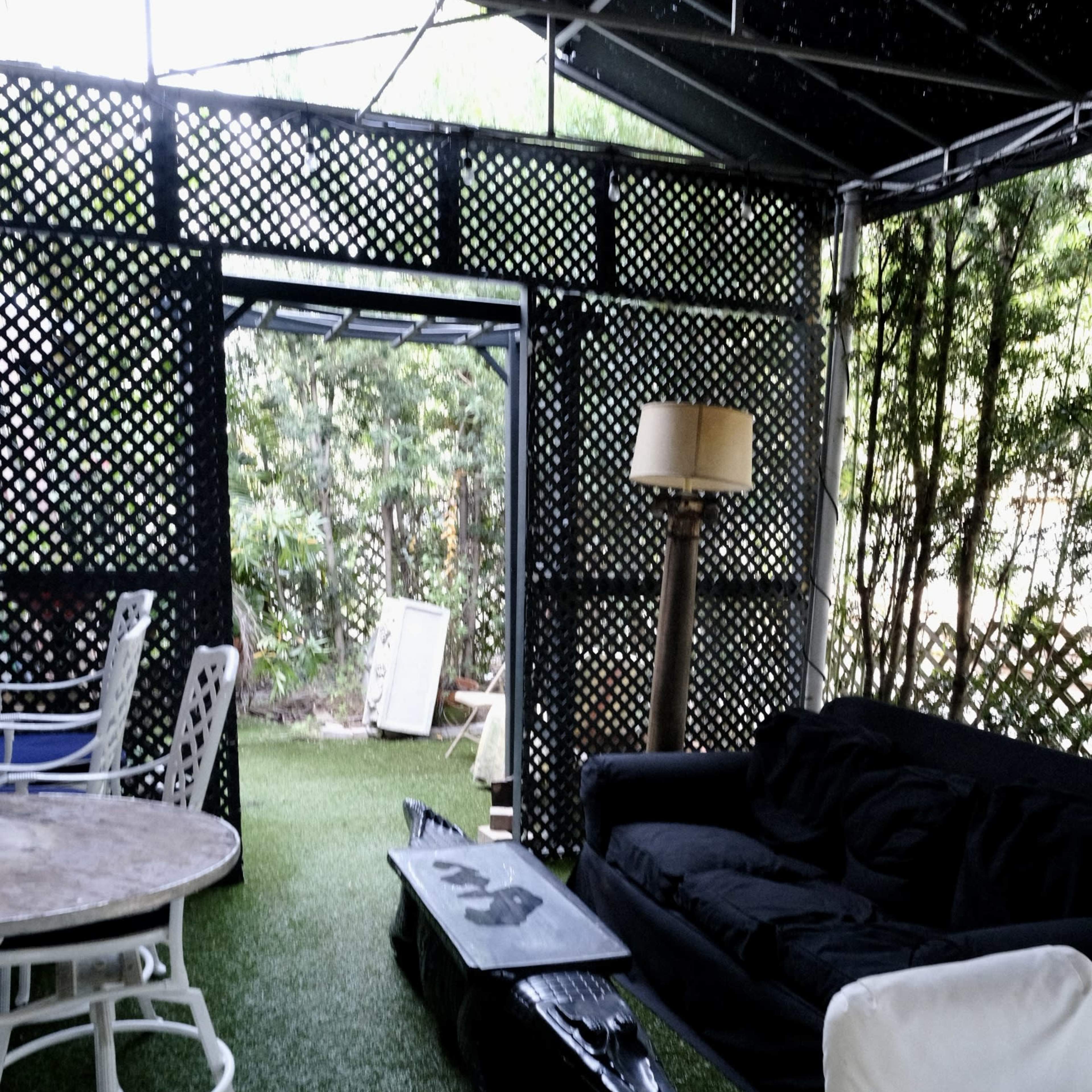 A cozy outdoor seating area with a black couch, a table on wheels, and a lamp, all surrounded by greenery and lattice panels.