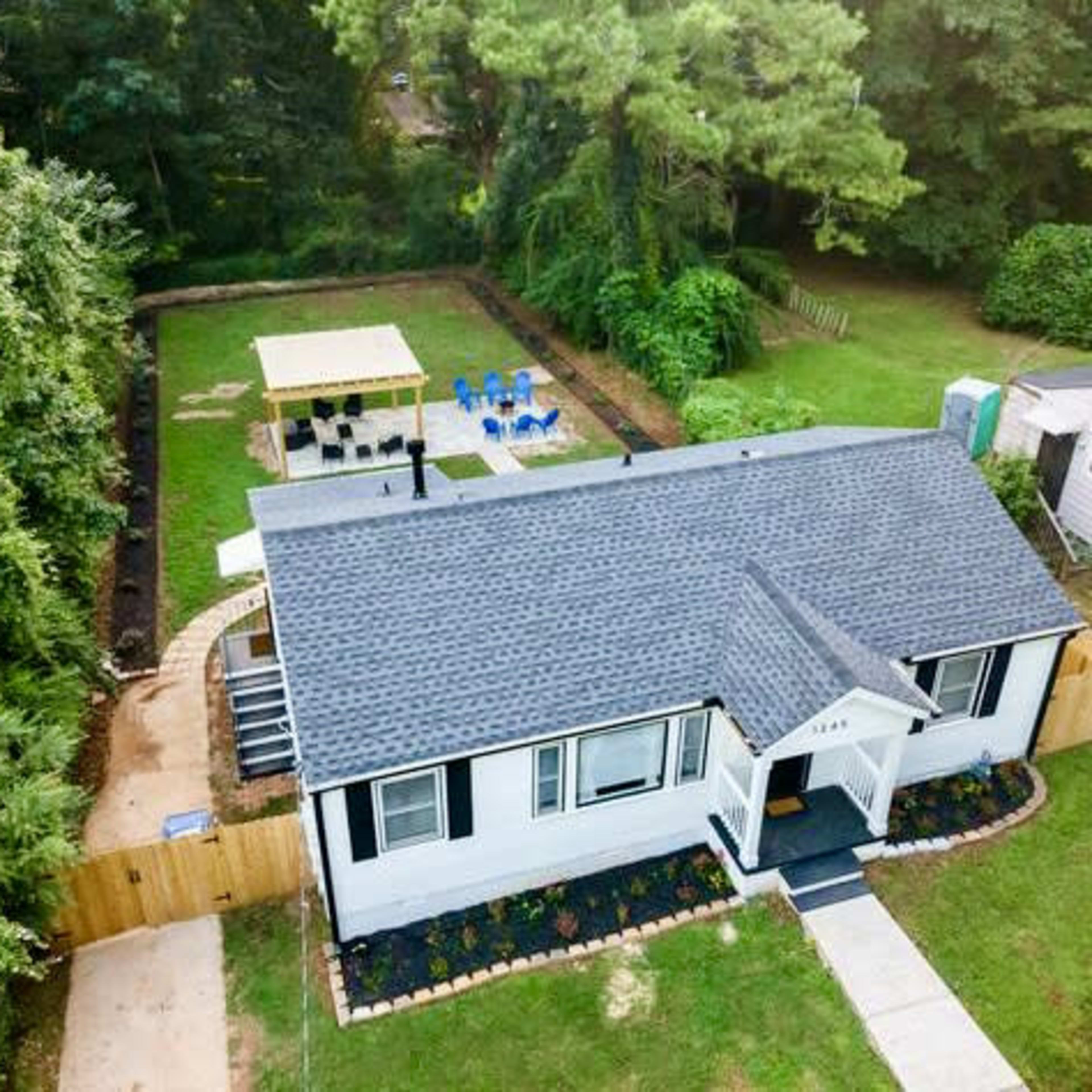 A single-story house with a black roof, surrounded by green foliage and featuring a backyard with a seating area and a small lawn.