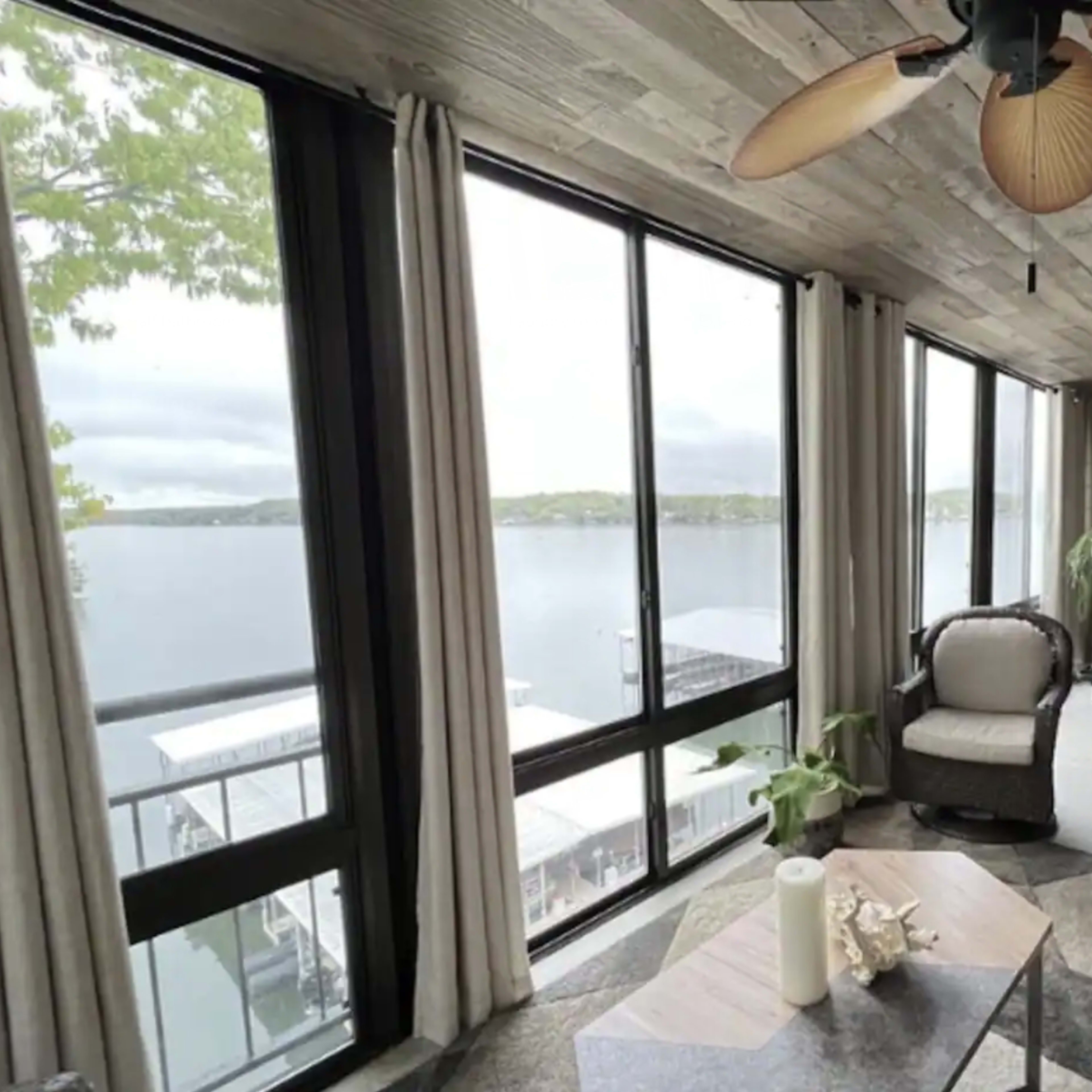 A sunroom with large windows overlooking a body of water, featuring comfortable seating, a coffee table, and a ceiling fan.