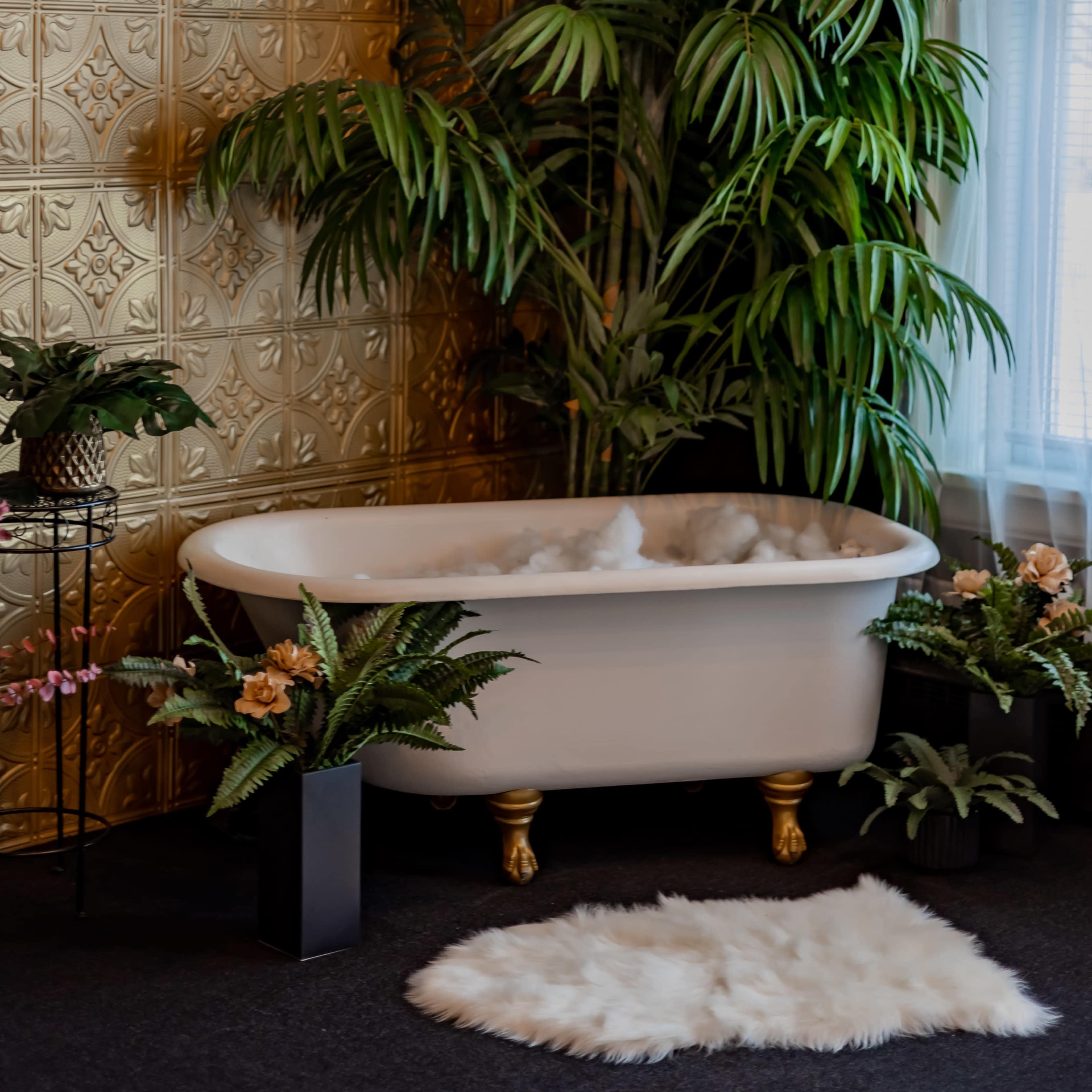 A freestanding white bathtub is surrounded by potted plants and decorative flowers against a gold-patterned wall.