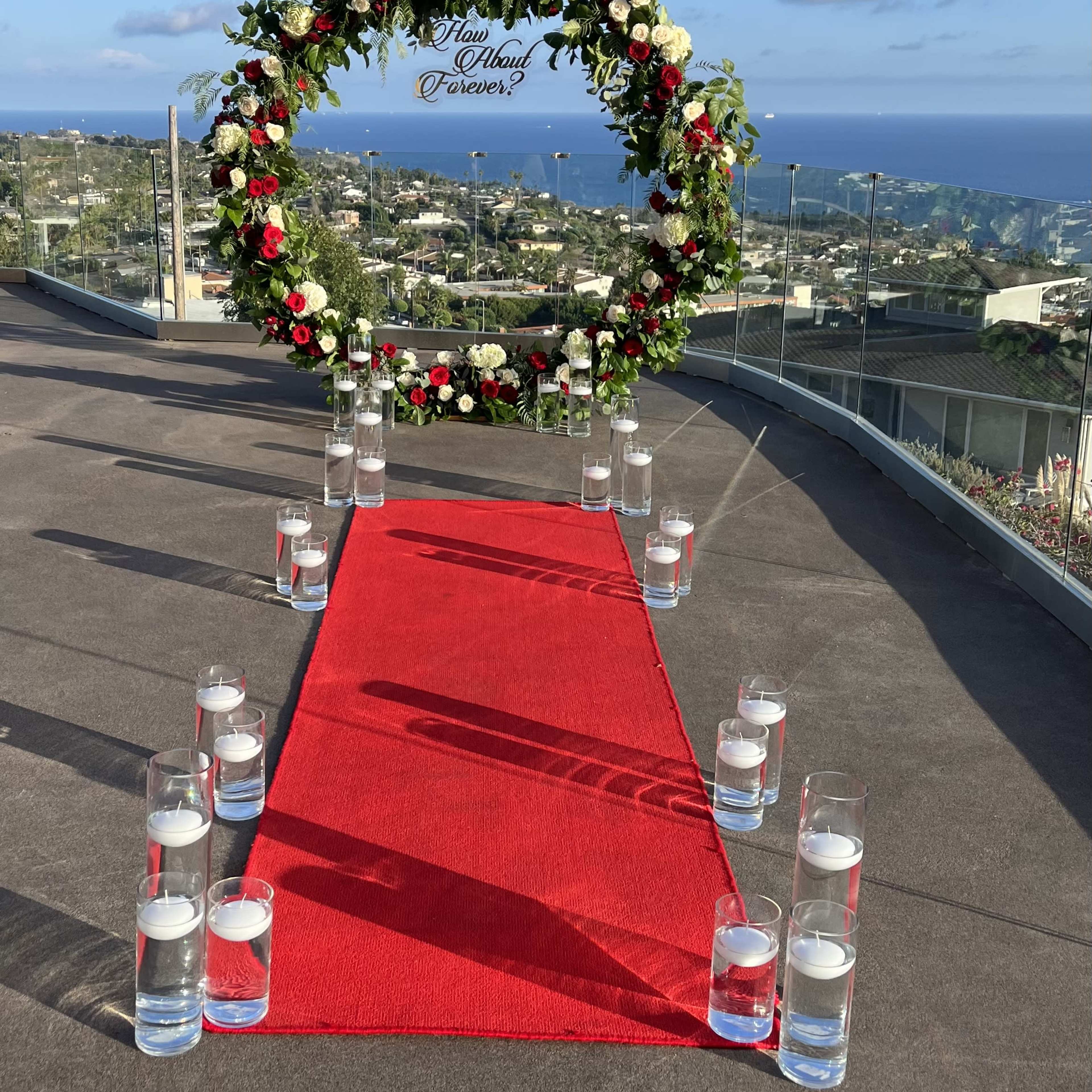 A circular floral arch adorned with red and white flowers stands at the end of a red carpet, flanked by glass candles, overlooking a panoramic view of a coastal landscape.