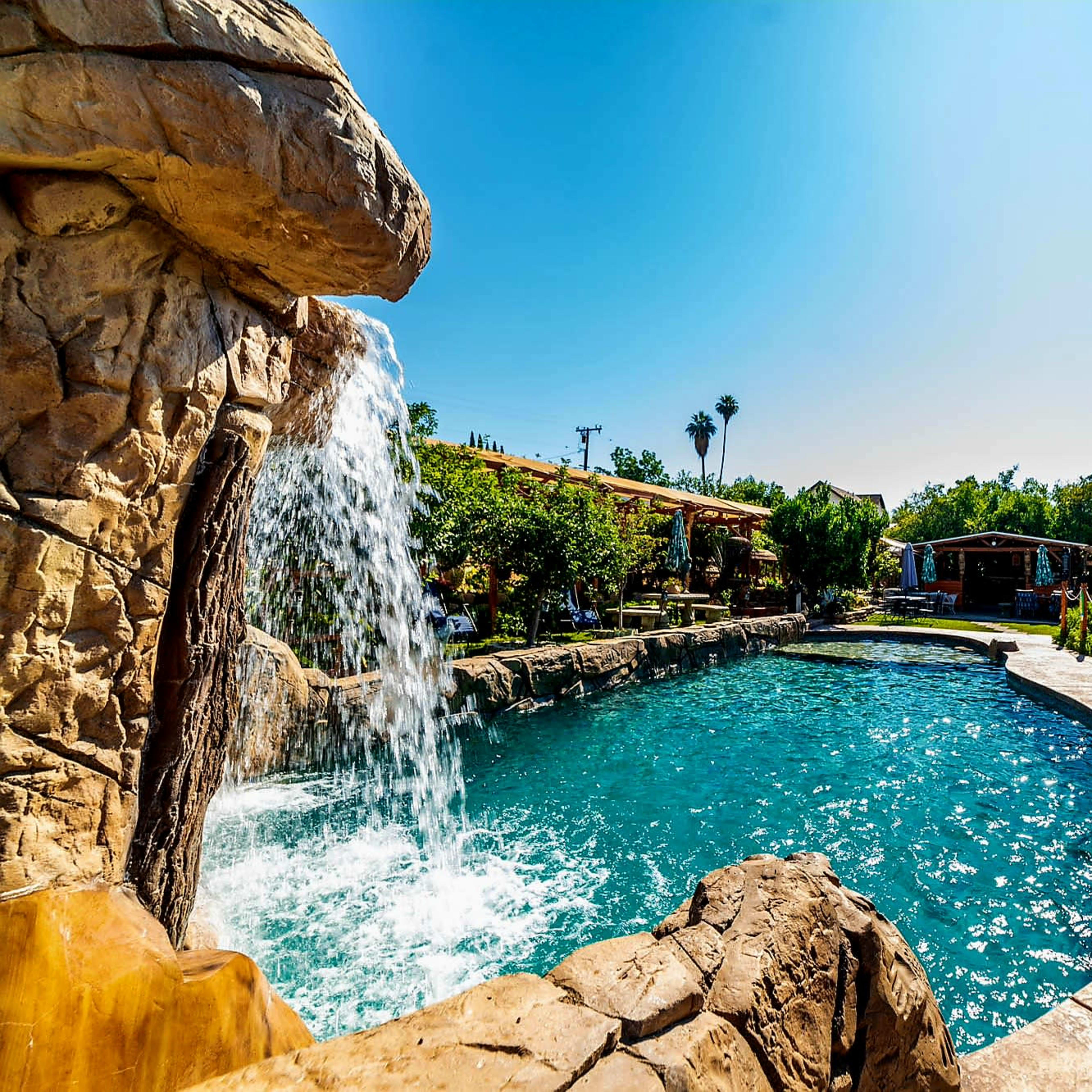 A pool surrounded by rock formations, a waterfall, and lush greenery under a clear blue sky.
