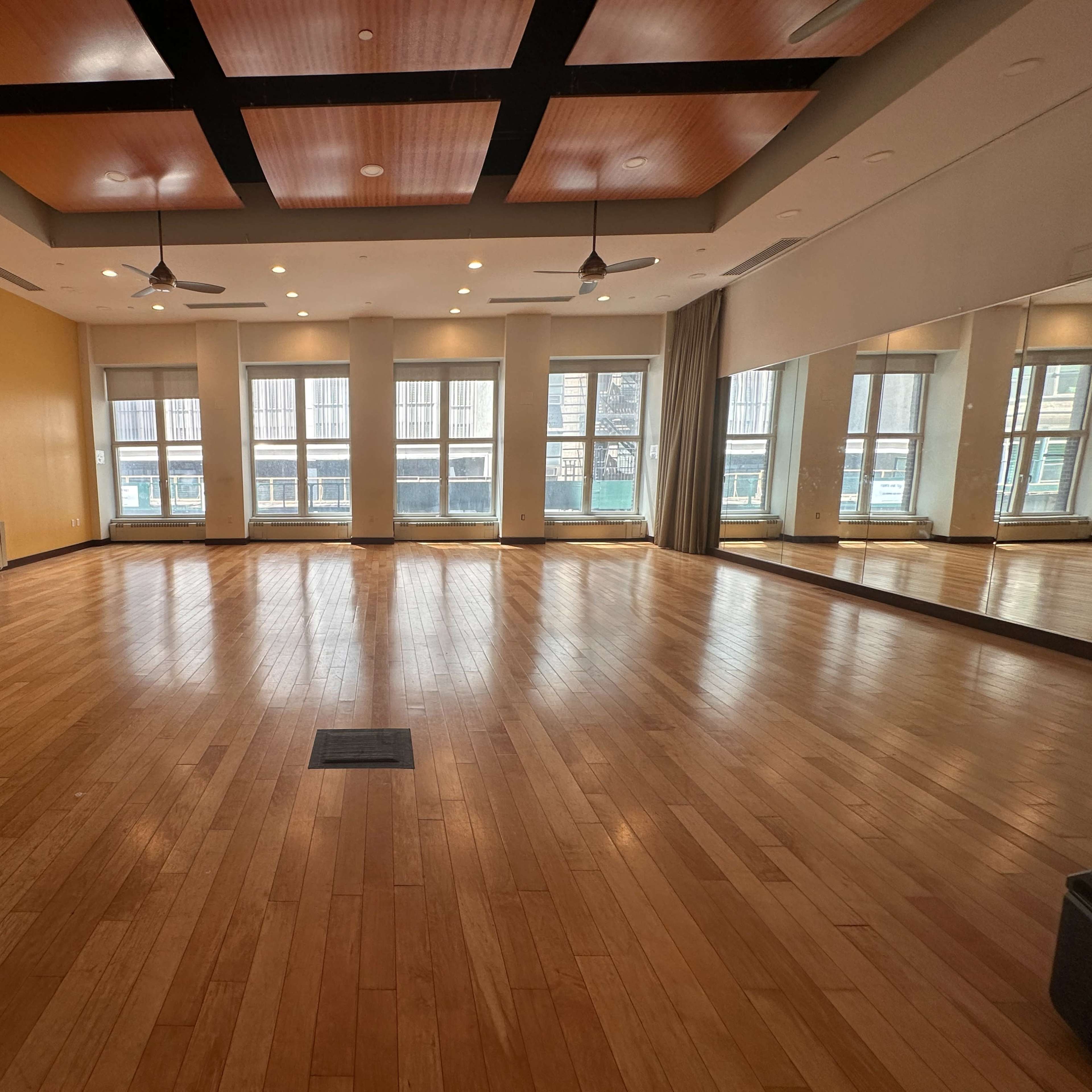 An empty wooden-floored room with large windows and a mirrored wall, featuring ceiling fans and a paneled ceiling design.