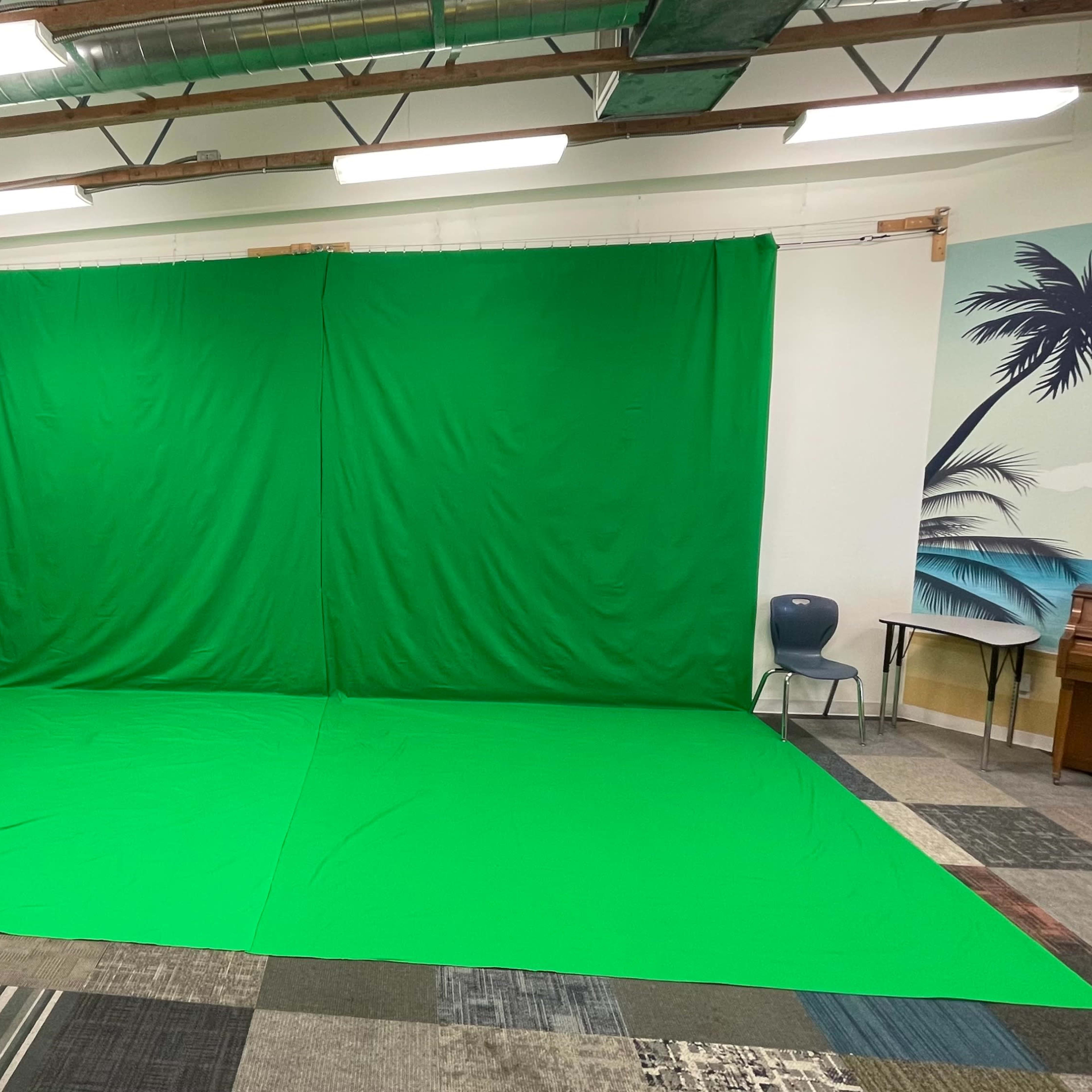 A room with a large green screen set up against the wall, accompanied by a piano, a chair, and a mural depicting palm trees and a beach.