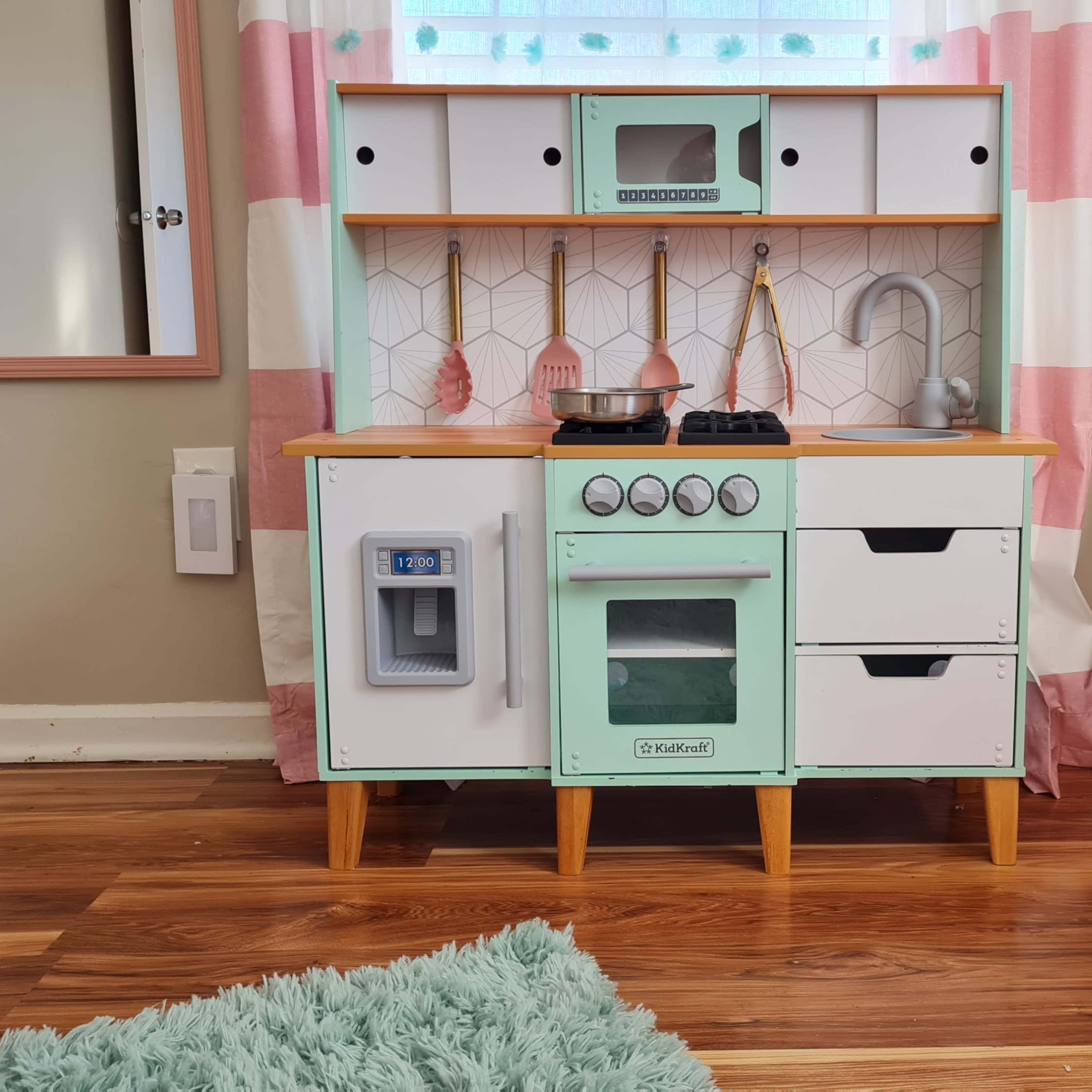 A children's play kitchen set with a mint green and white design, featuring a stove, sink, and storage drawers, positioned on a hardwood floor.