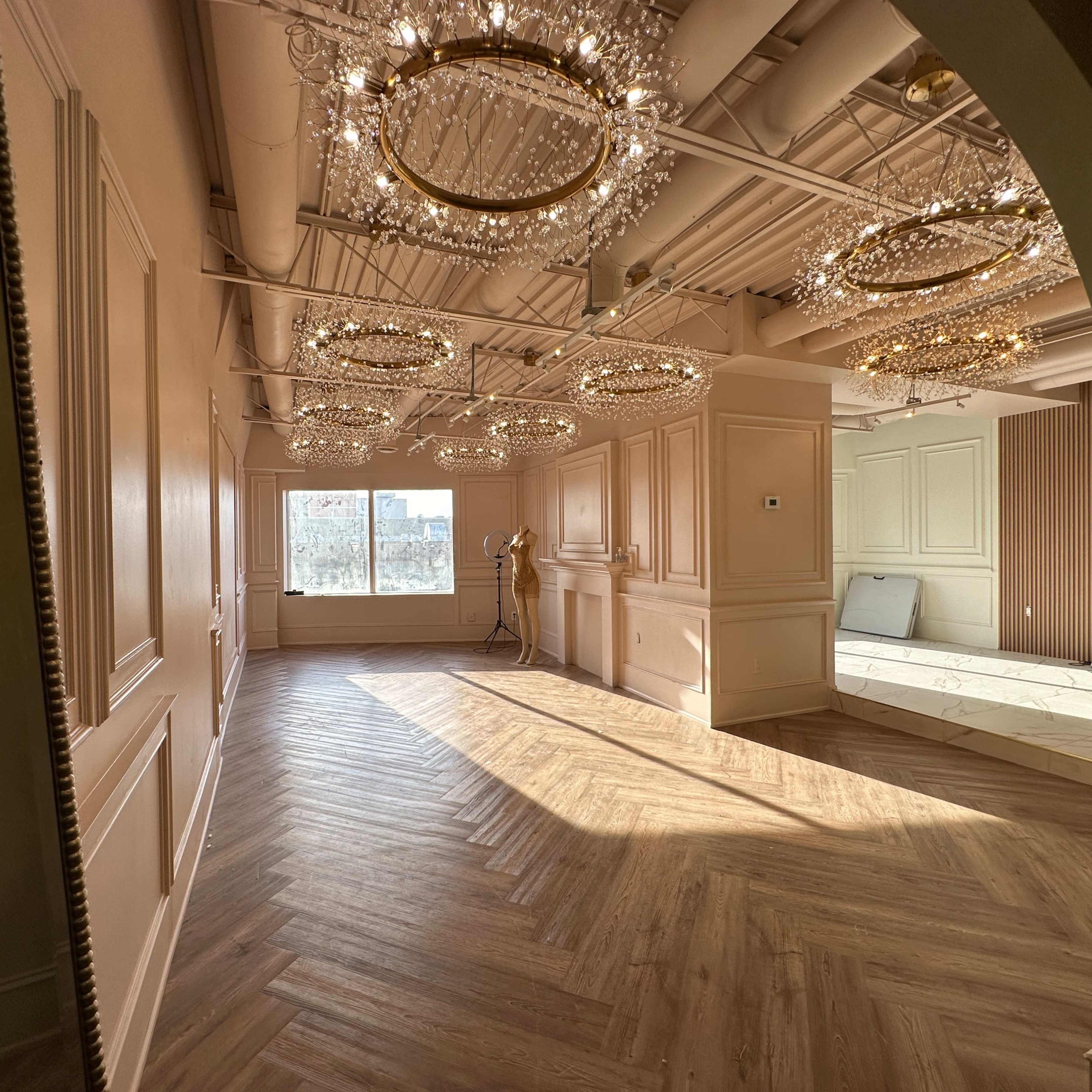 A spacious room with herringbone wooden flooring, featuring several ornate chandeliers and large windows allowing natural light to enter.