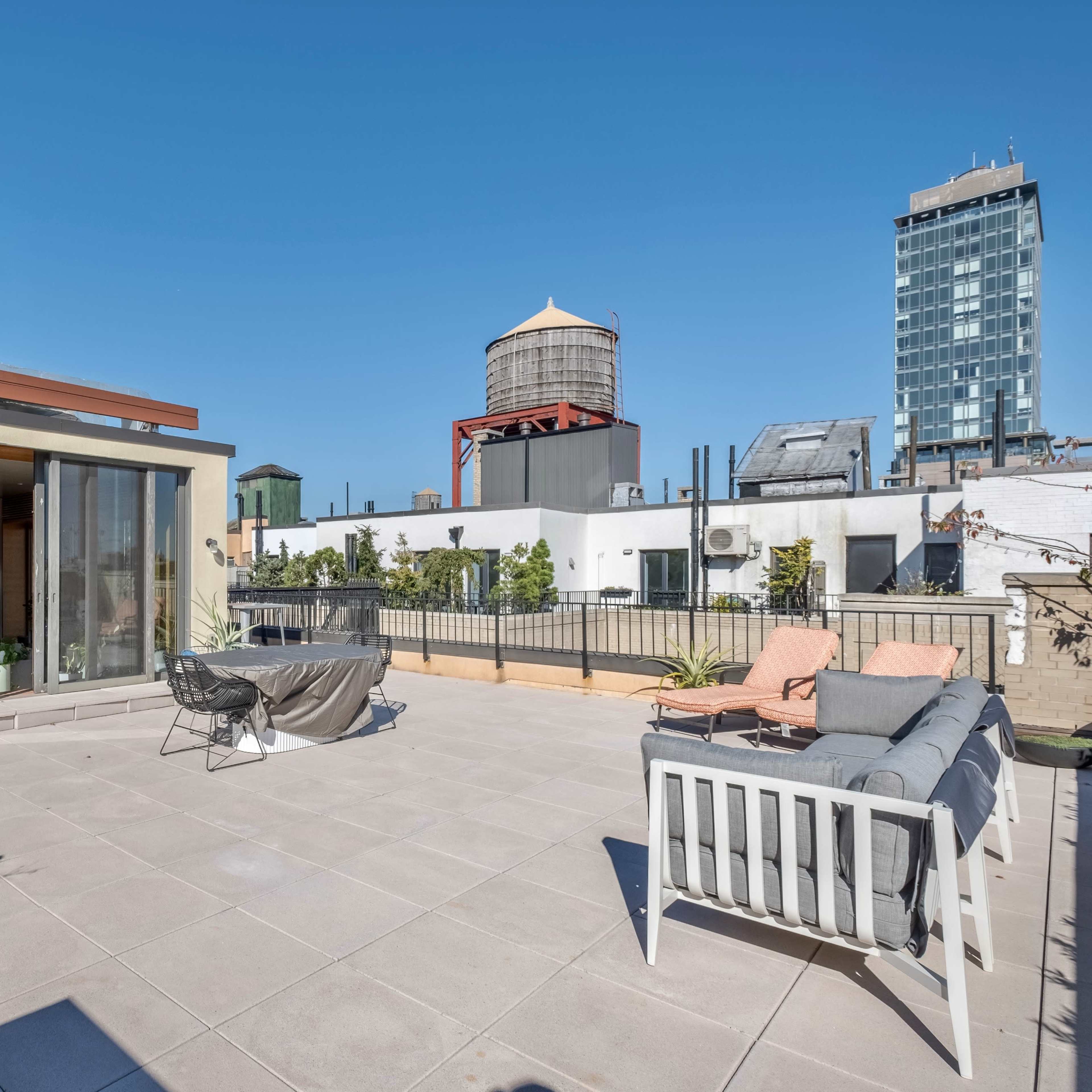 A rooftop terrace with seating areas, potted plants, and several water towers in the background against a clear blue sky.