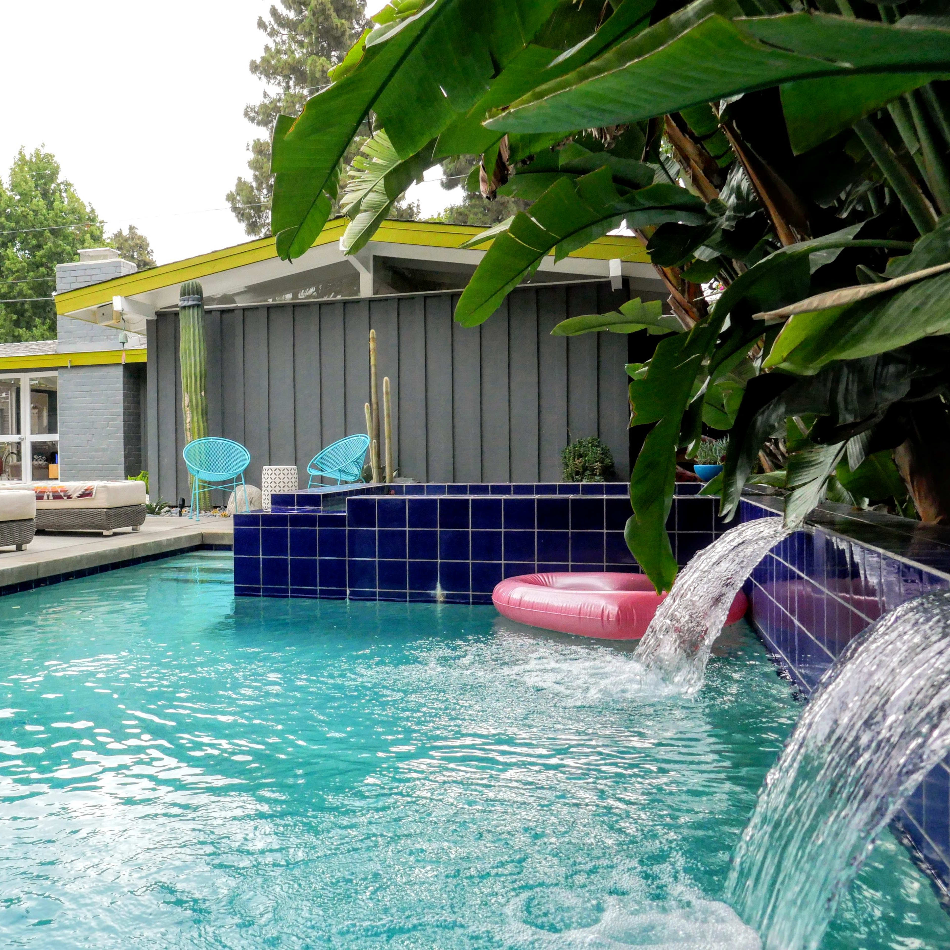 A backyard with a swimming pool, a waterfall feature, lounge furniture, and palm trees against a modern home facade.