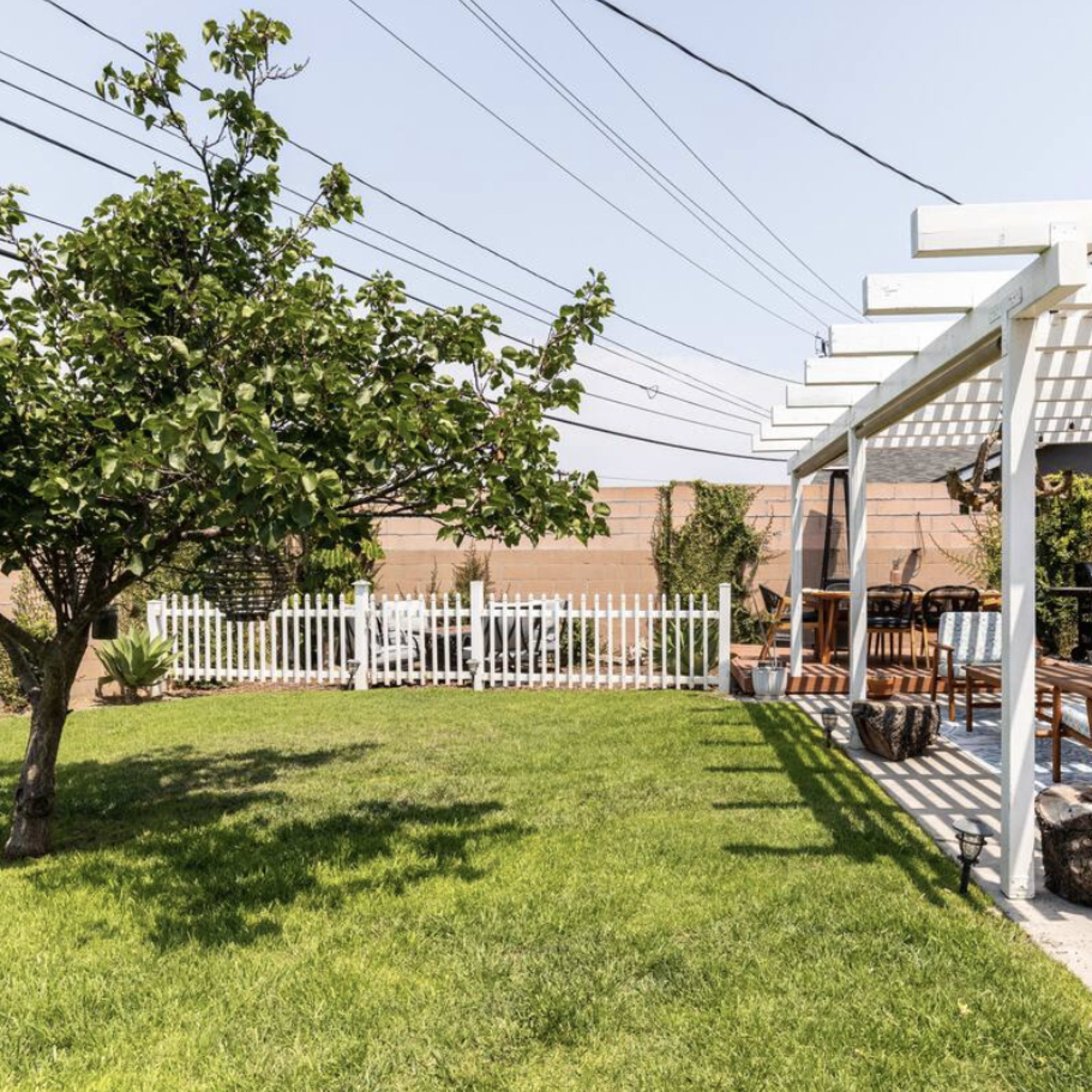A backyard with a green lawn, a white pergola, a seating area, and a small tree.