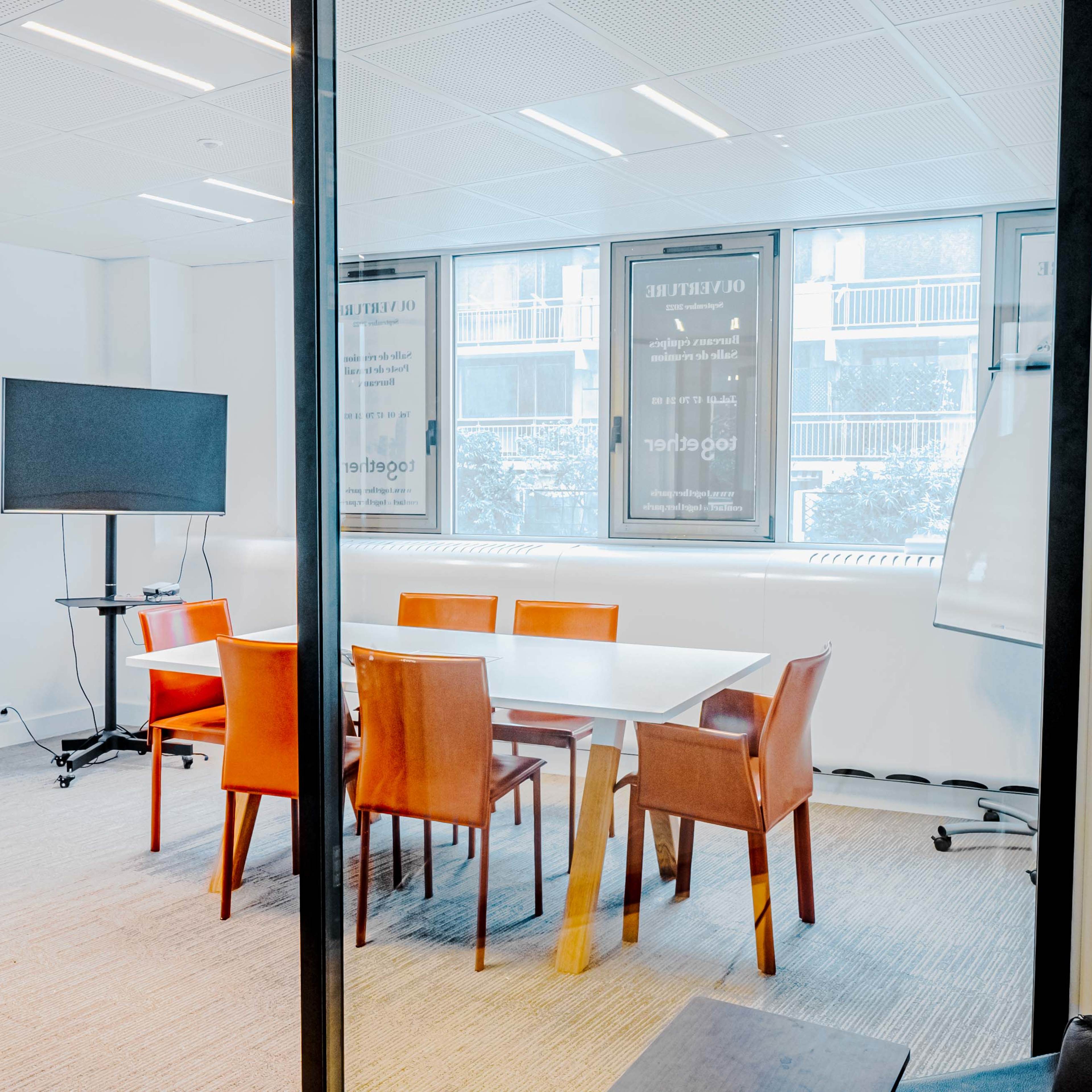 A glass-walled conference room features a white table surrounded by orange chairs, with a television and a whiteboard positioned nearby.