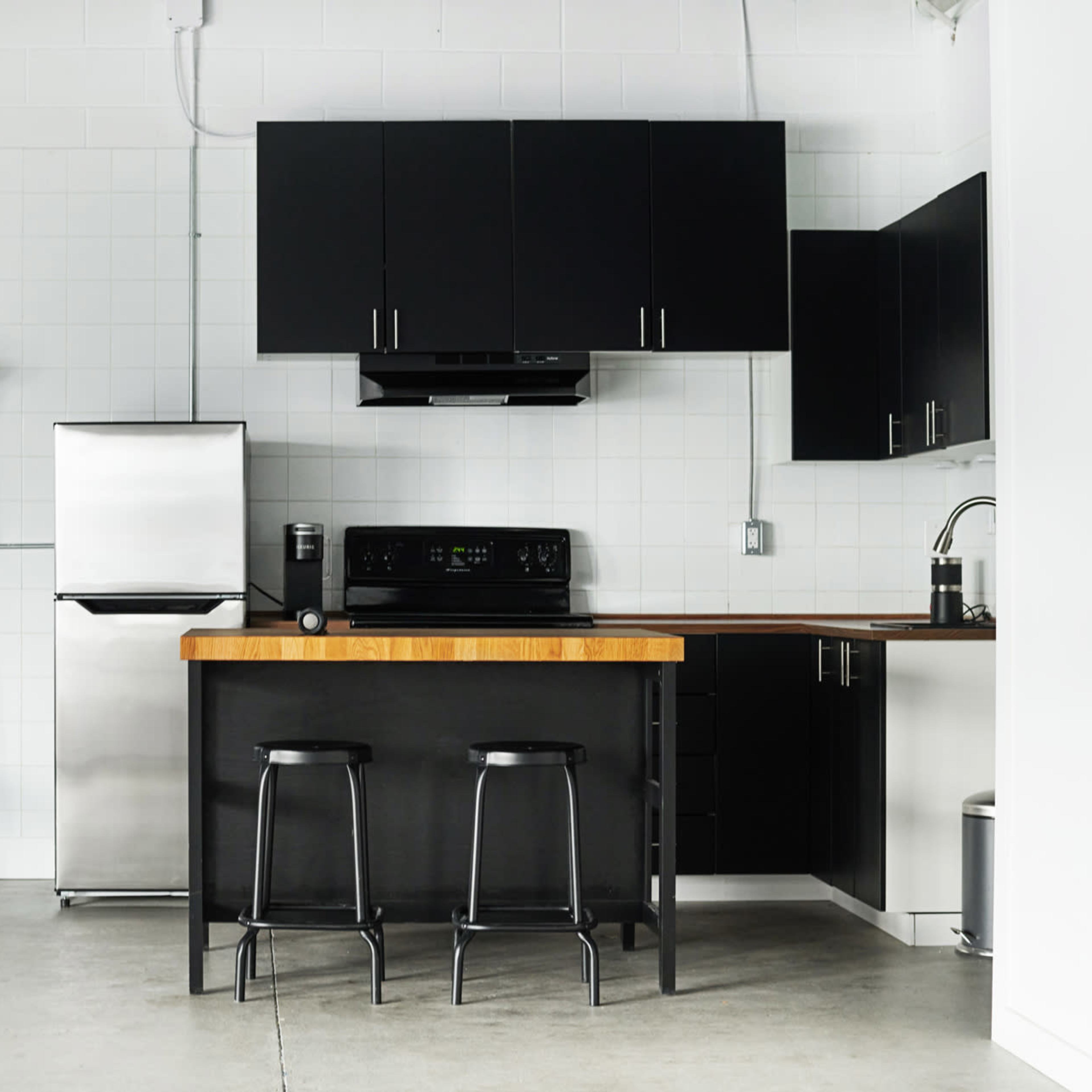 A modern kitchen with a stainless steel refrigerator, black cabinetry, a wooden countertop with two stools, and a black sofa with a red pillow.