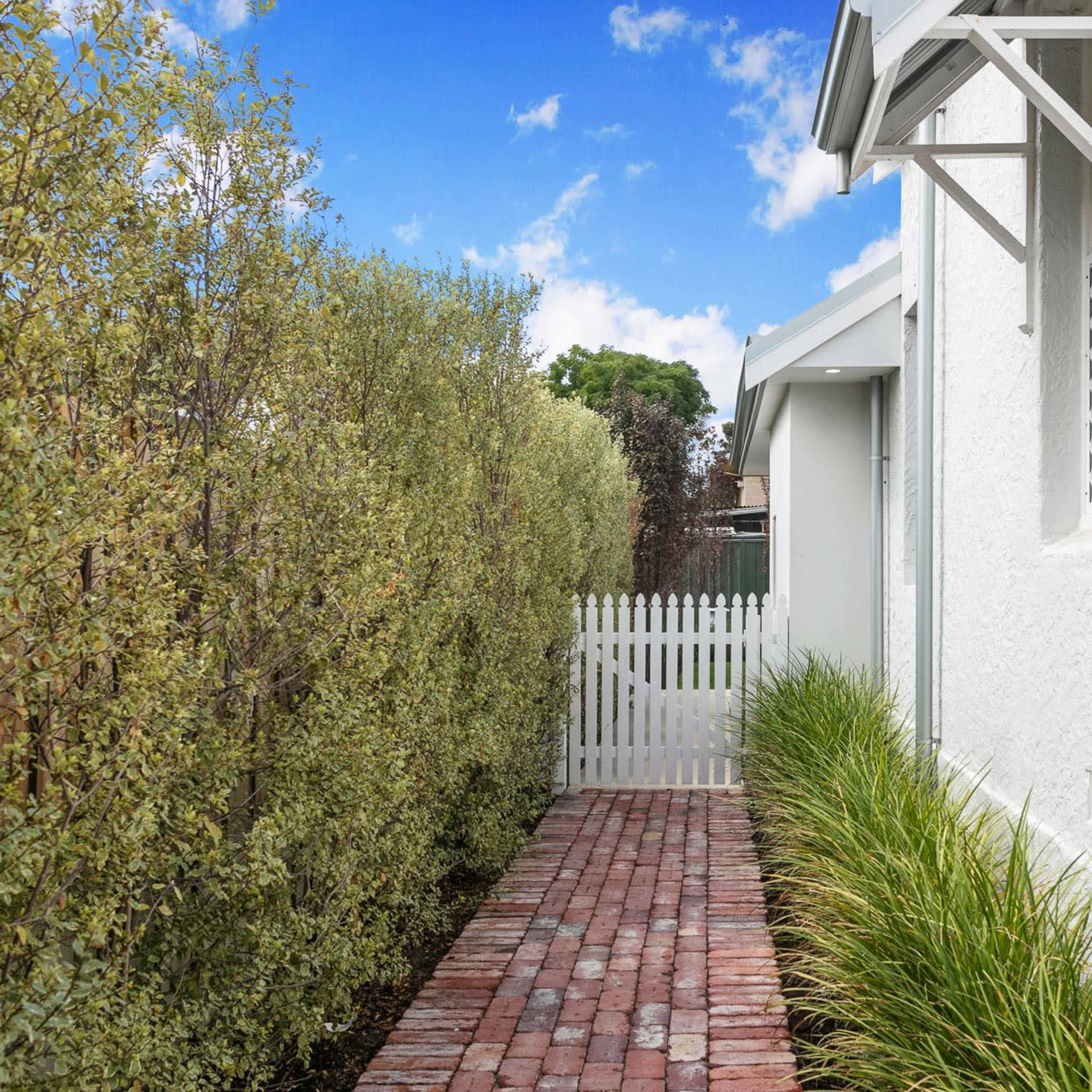 A narrow brick pathway lined with tall greenery leads to a white home, with a wooden picket fence visible at the entrance.