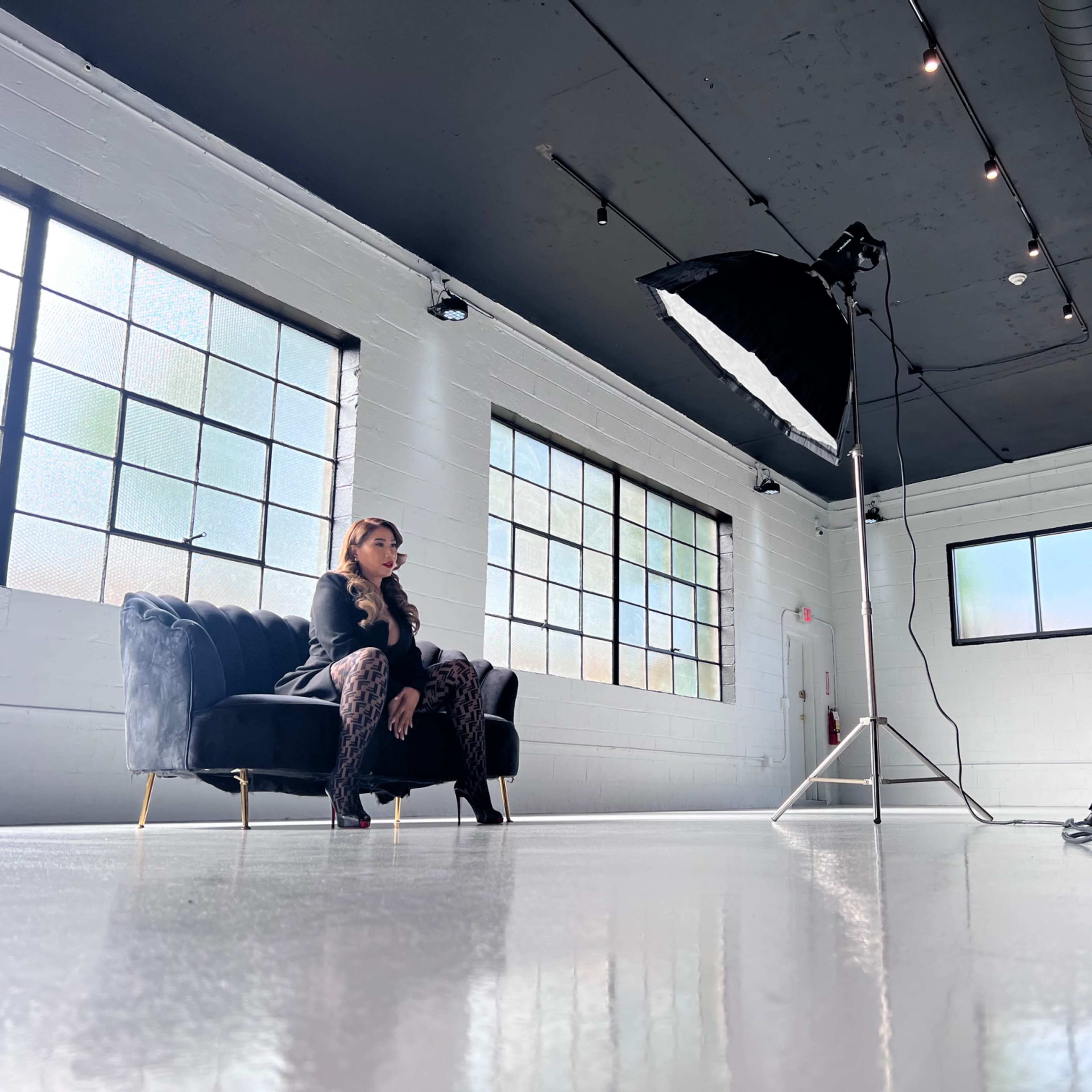 A photographer captures a model sitting on a black couch in a large, well-lit studio space.
